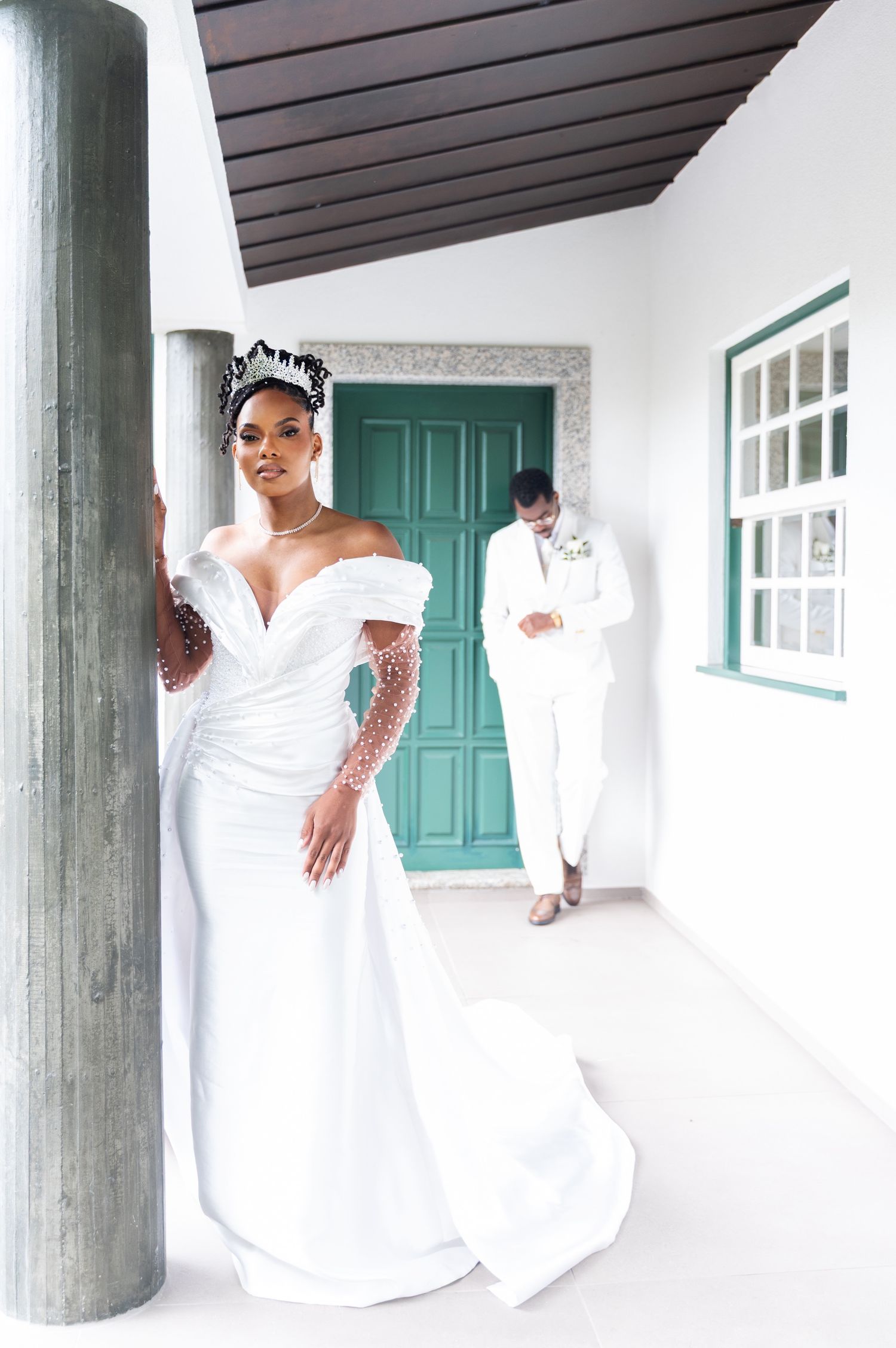 Elegant bride in fitted white pearl wedding dress with tiara poses by pillar, groom in background.