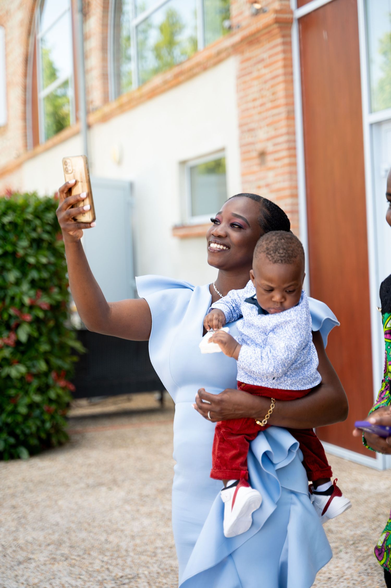 Smiling woman in blue dress takes selfie while holding baby outdoors near brick building.
