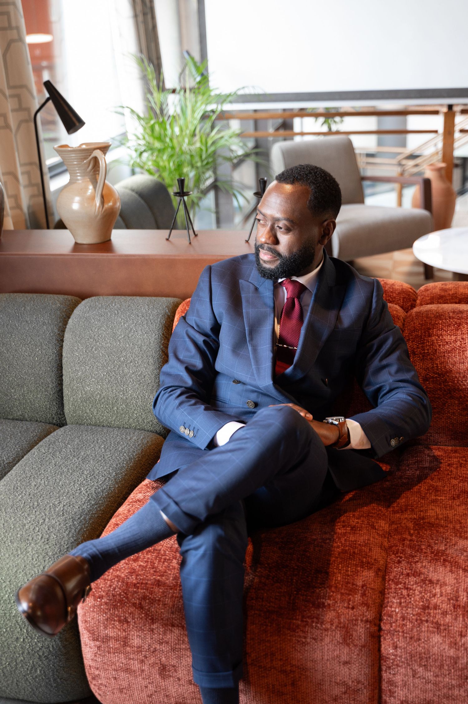 Elegant Black man in navy blue suit with burgundy tie sitting relaxed on a rust-colored velvet sofa in modern lounge.