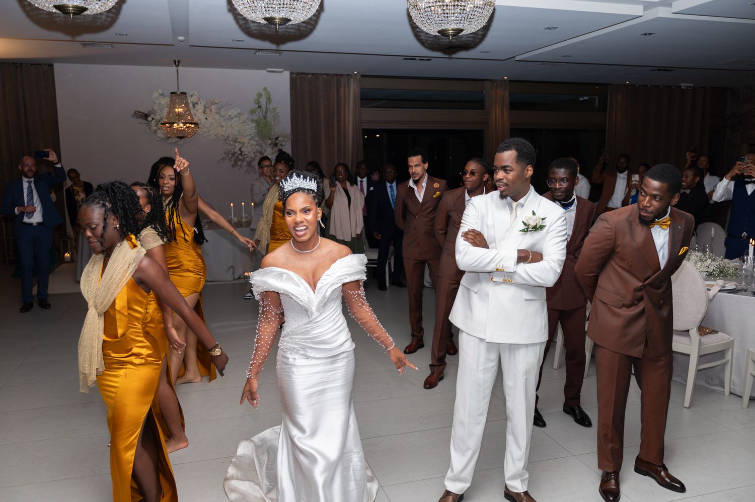 Bride in white gown and crown dances joyfully with wedding party in golden bridesmaid dresses at reception.