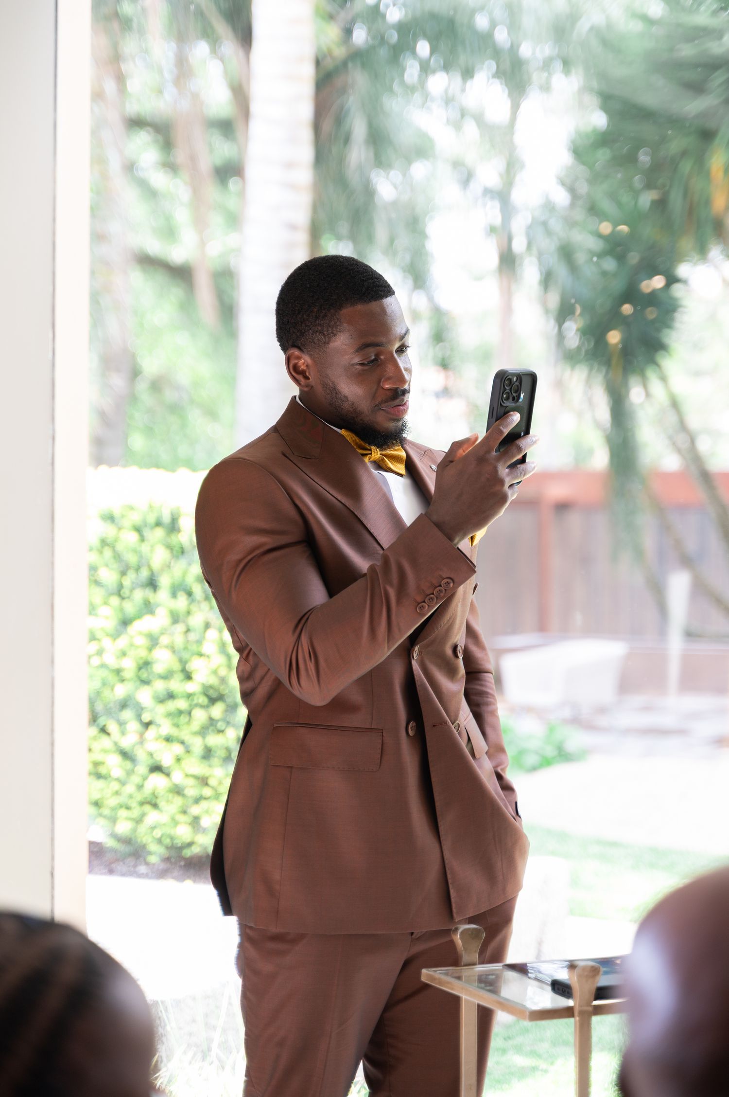 Stylish wedding guest in brown suit with gold bow tie photographs ceremony on smartphone.