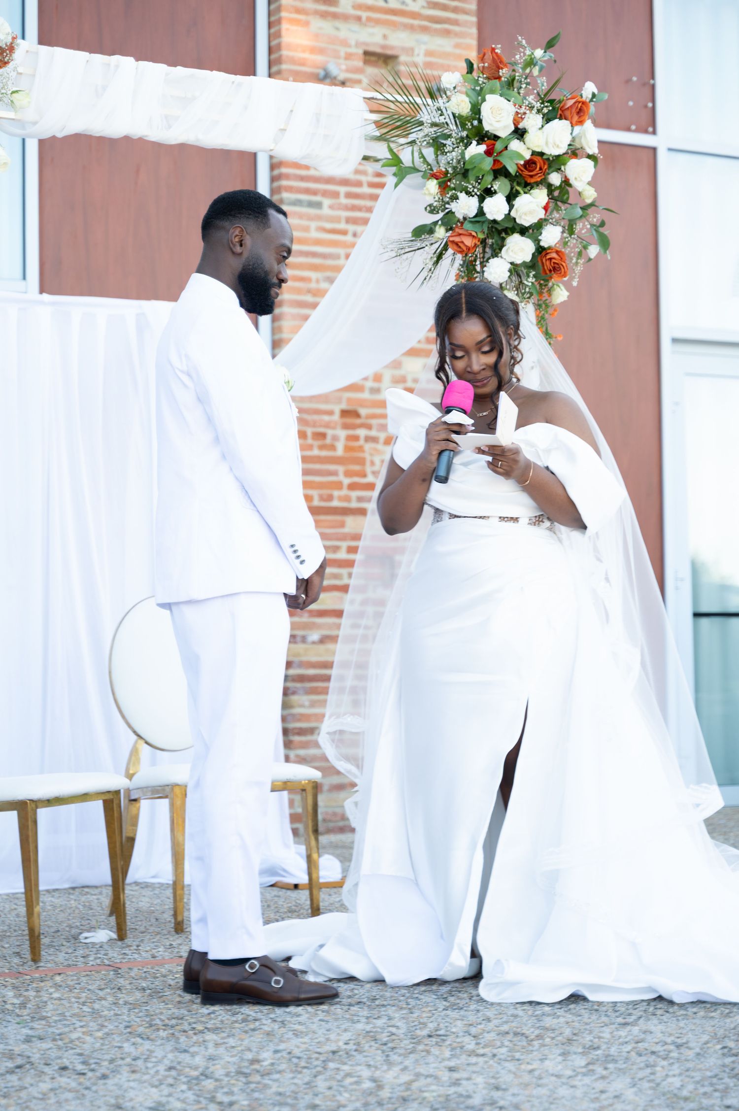 Bride in white off-shoulder gown reads vows to groom under floral wedding arch.
