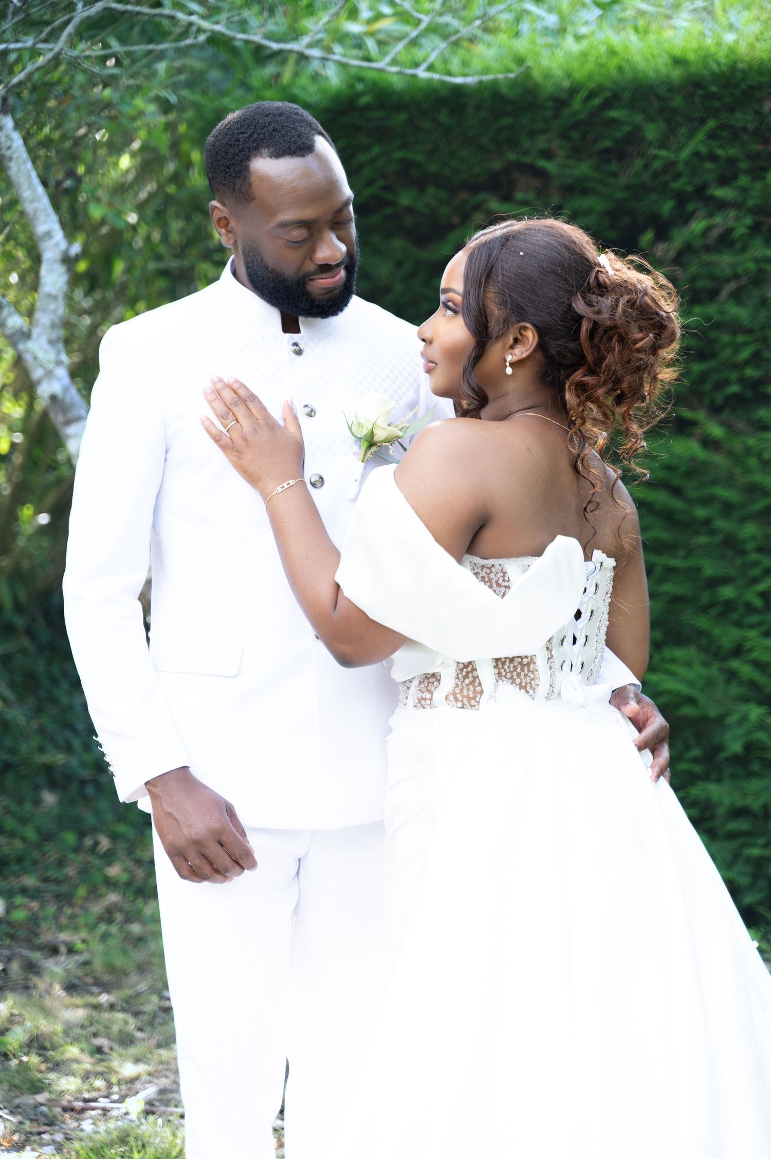Elegant couple in matching white wedding attire sharing a tender moment in a lush garden setting.