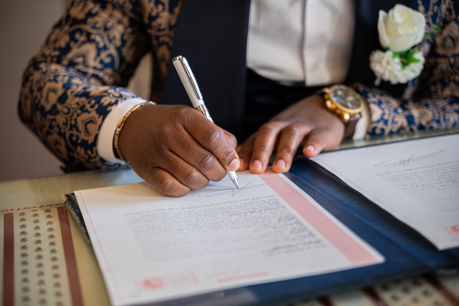Close-up of groom signing official wedding documents with pen wearing decorative suit and gold watch.
