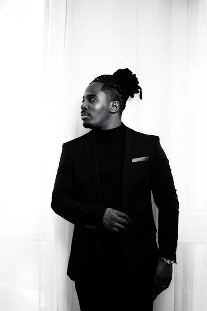 Black and white portrait of a stylish man in a black suit with dreadlocks, posing confidently against a white wall.