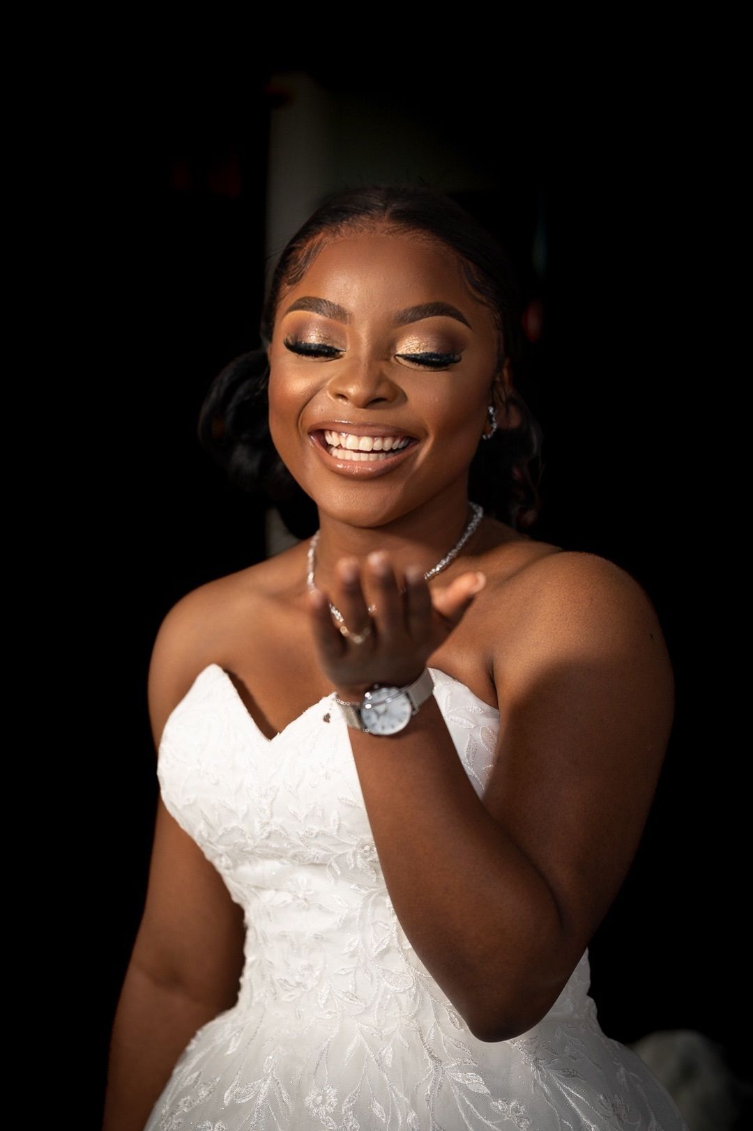 Radiant bride in strapless white lace gown smiling joyfully while blowing a kiss against a dark background.