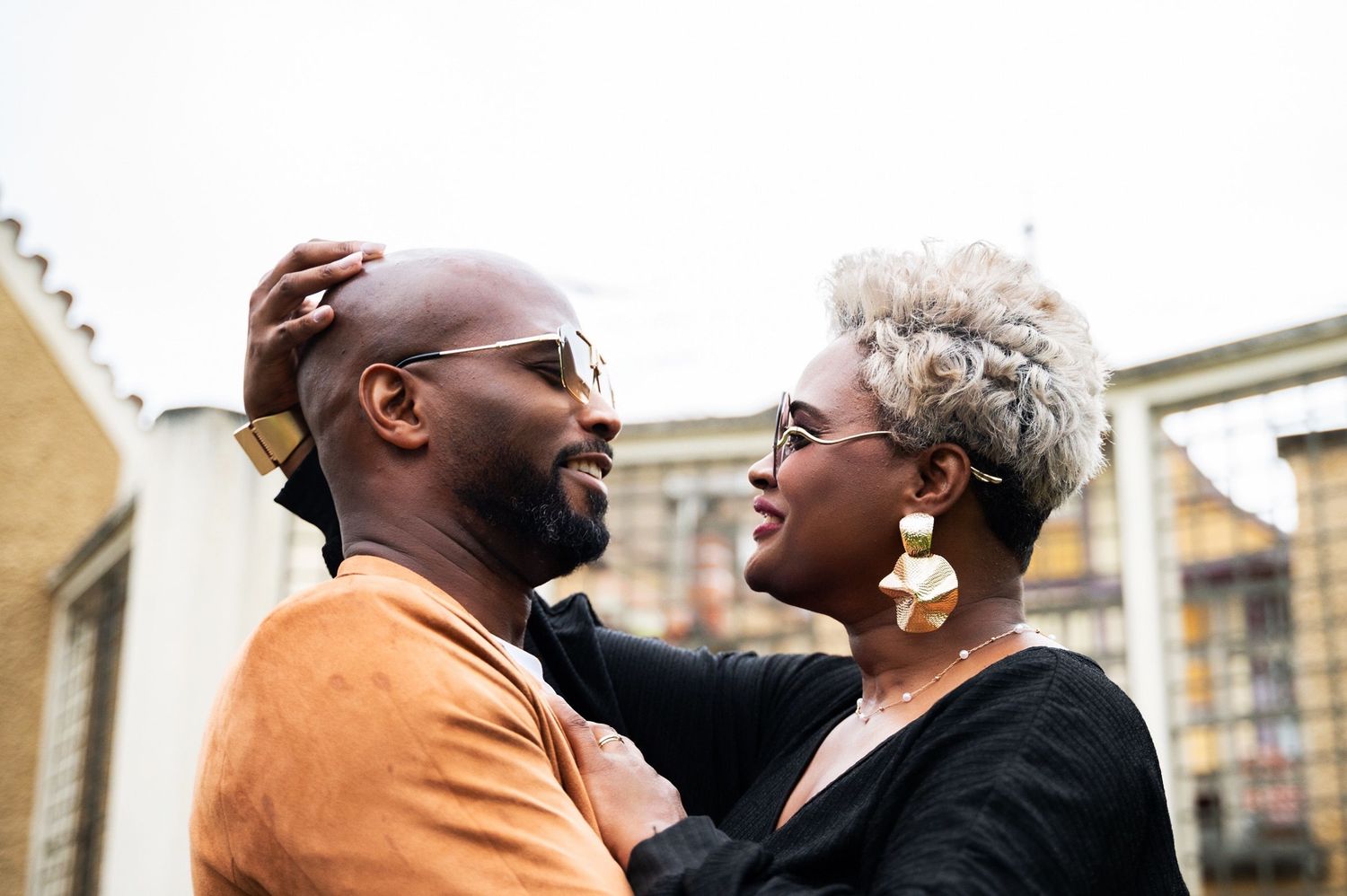 Black couple smiling and embracing outdoors, woman touching mans head affectionately.