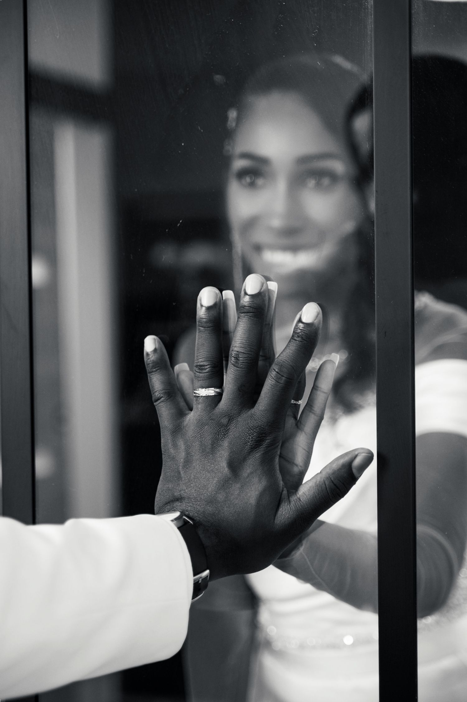 Groom places hand on glass door as smiling bride with wedding ring reflects in black and white photo.