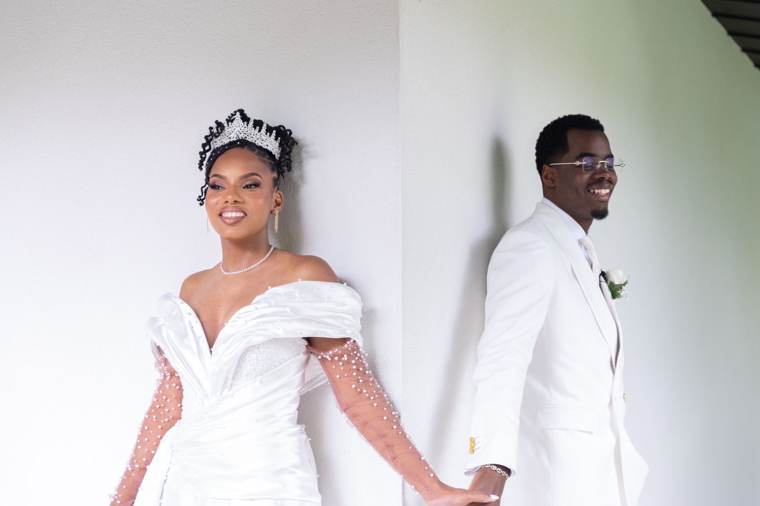Smiling Black bride and groom in matching white wedding attire holding hands at outdoor venue.