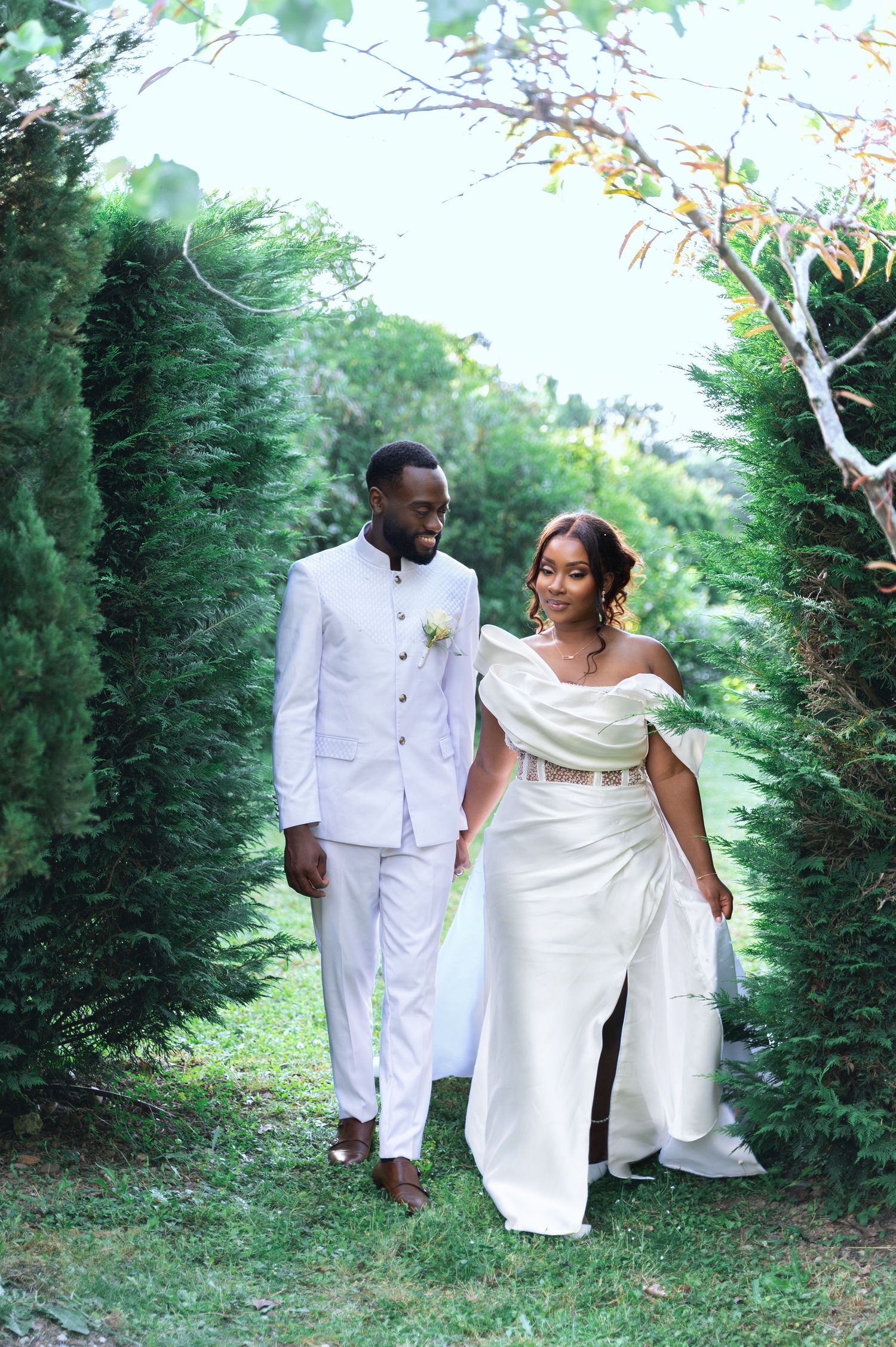 Black bride and groom in matching white wedding attire walking hand in hand through a green tree-lined garden path.