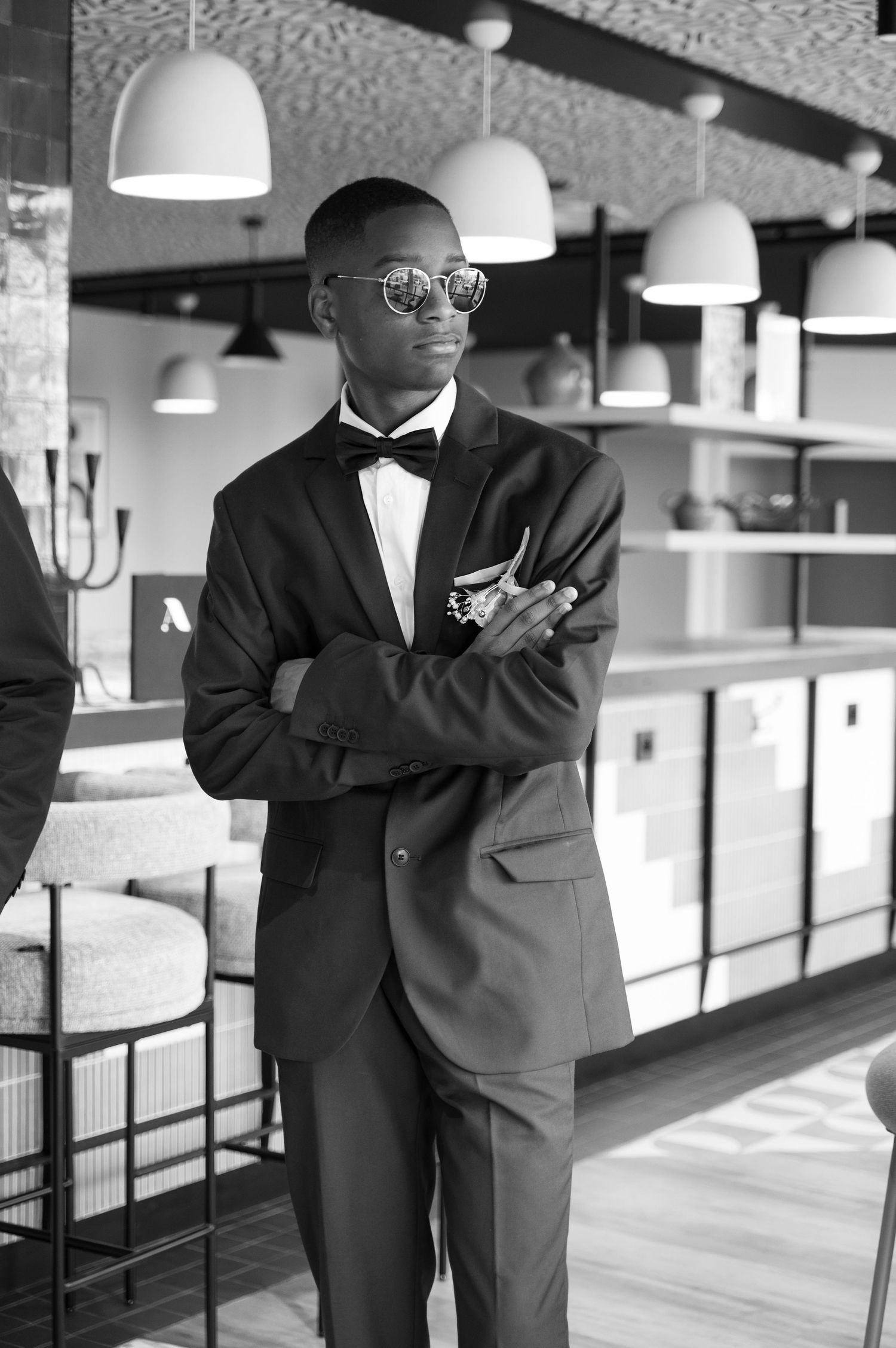 Stylish young Black man in black tuxedo with bow tie and round sunglasses standing with arms crossed in modern venue.