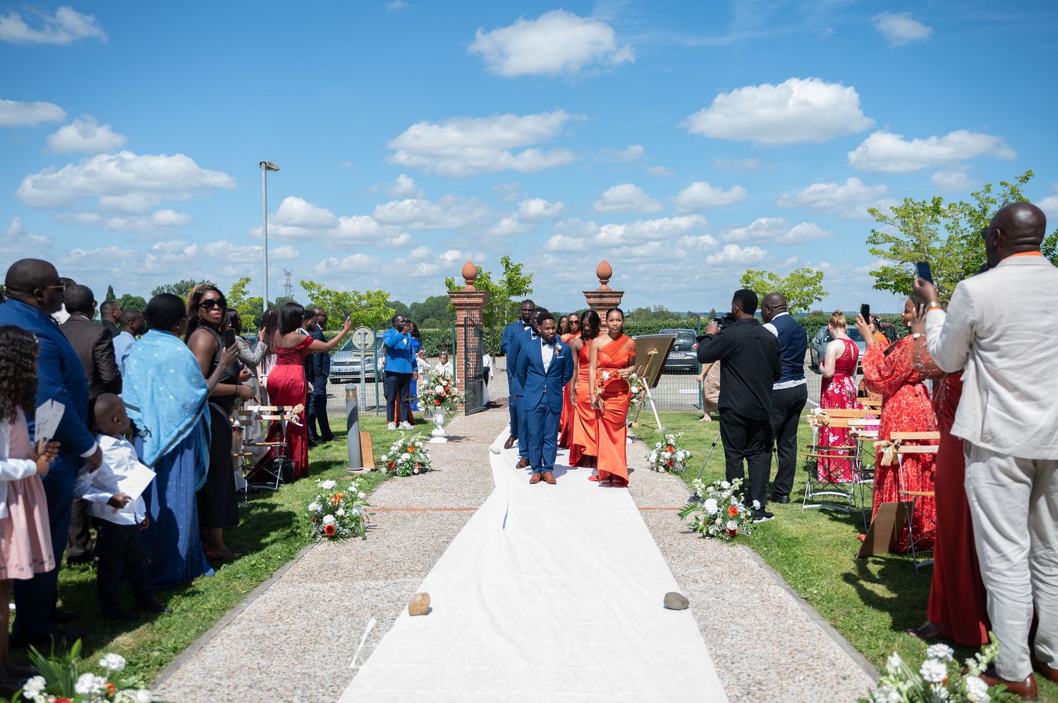 Outdoor wedding ceremony procession on white aisle runner with guests watching on sunny day.