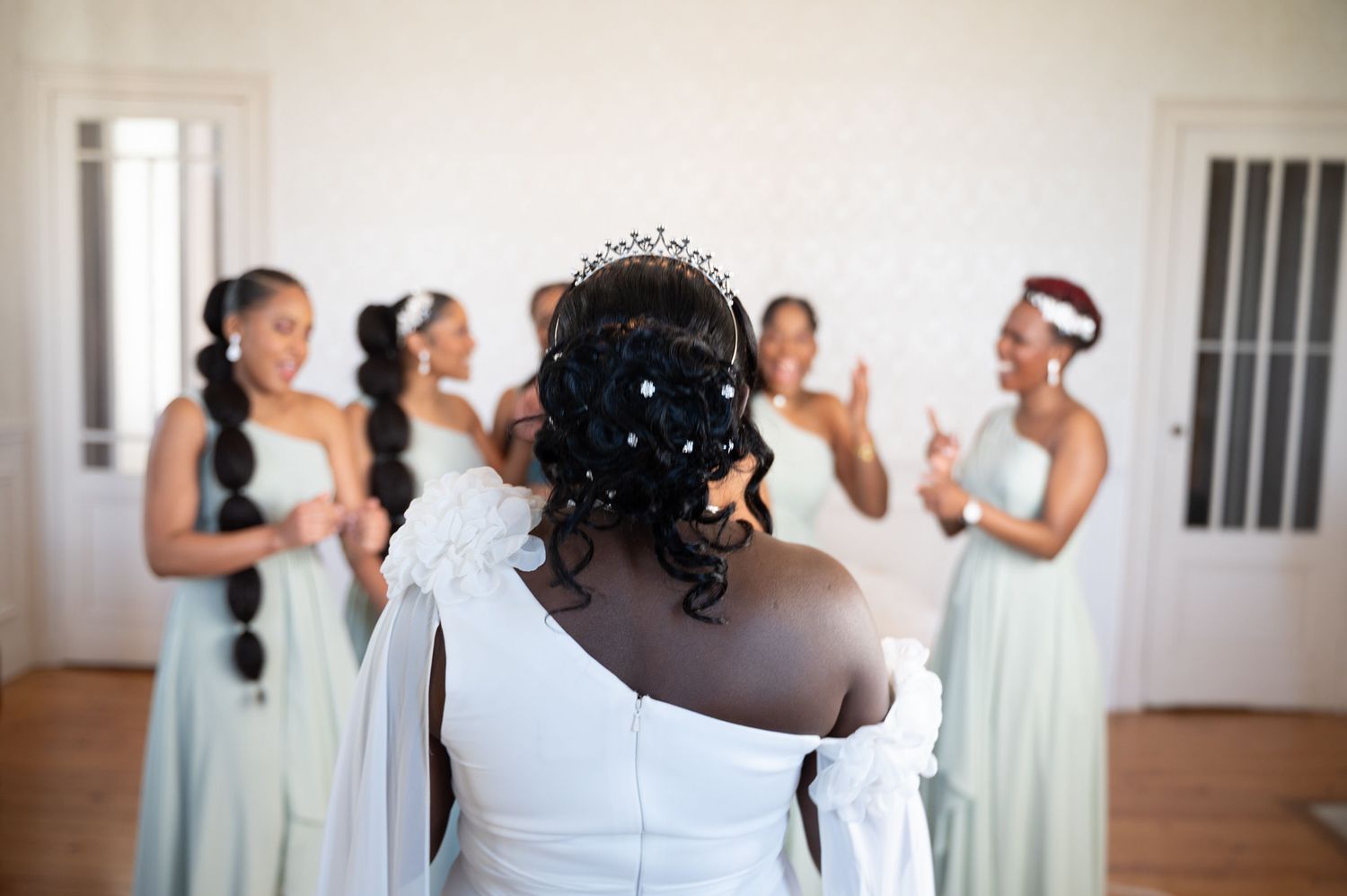 Bride in white gown and tiara faces her bridesmaids in mint dresses during a wedding preparation moment.