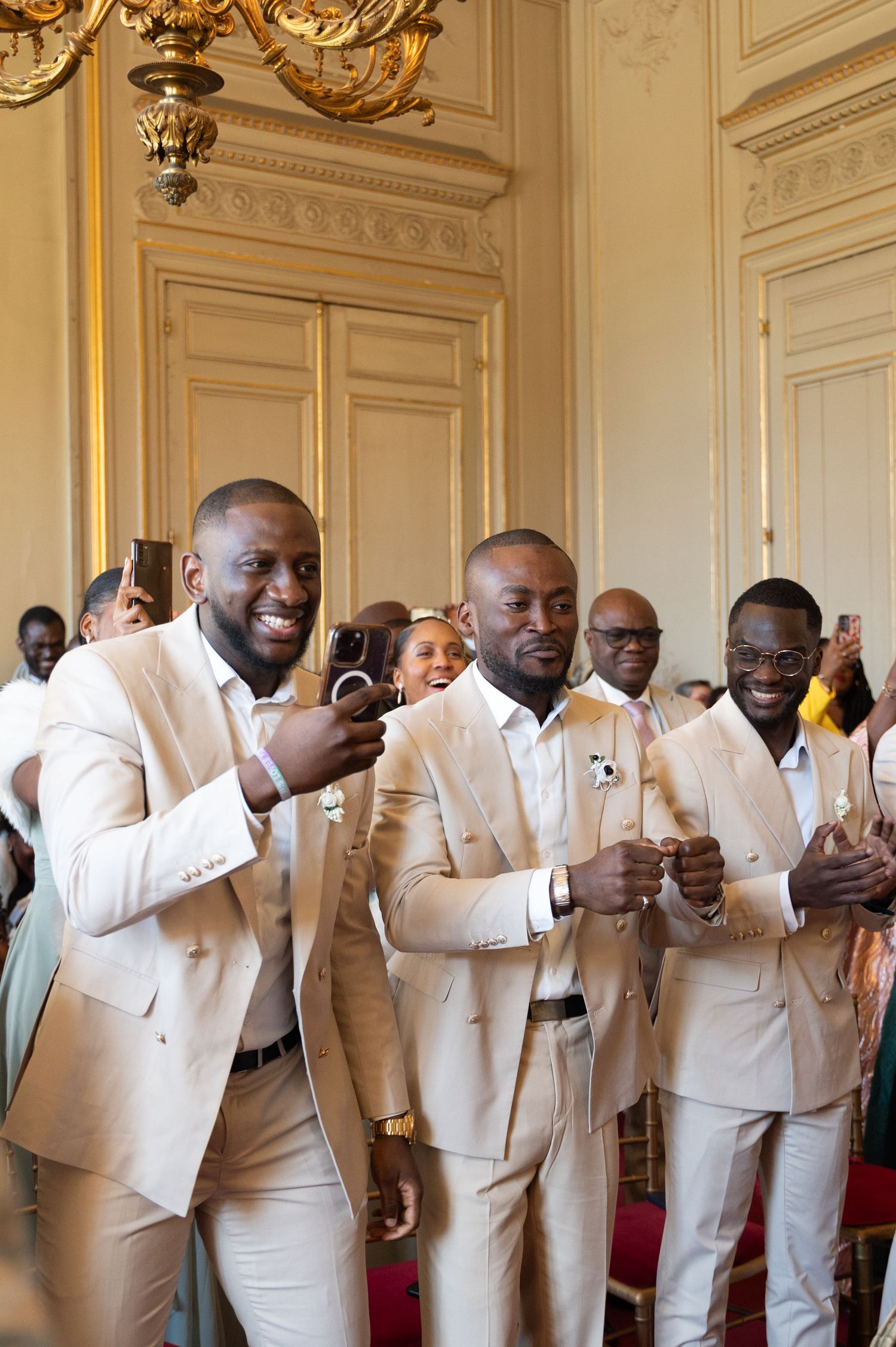 Groomsmen in matching champagne suits smile and clap during a wedding ceremony in an elegant ballroom.