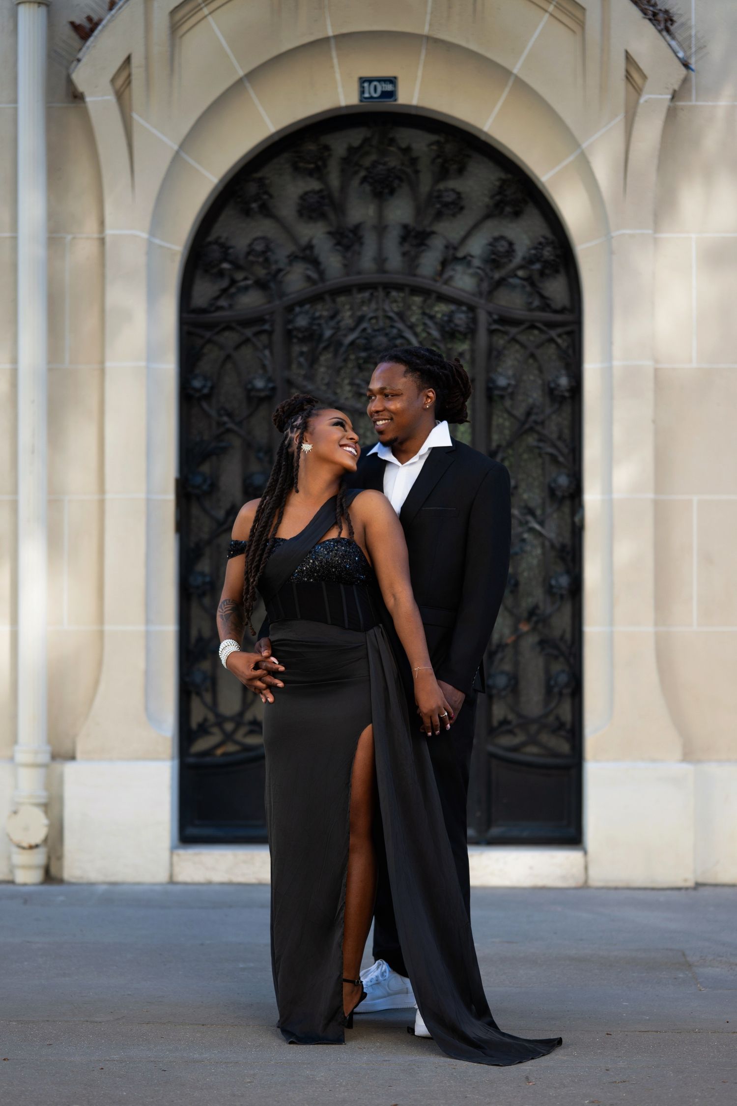 Elegant Black couple in matching black formal attire posing before ornate arched iron gate.