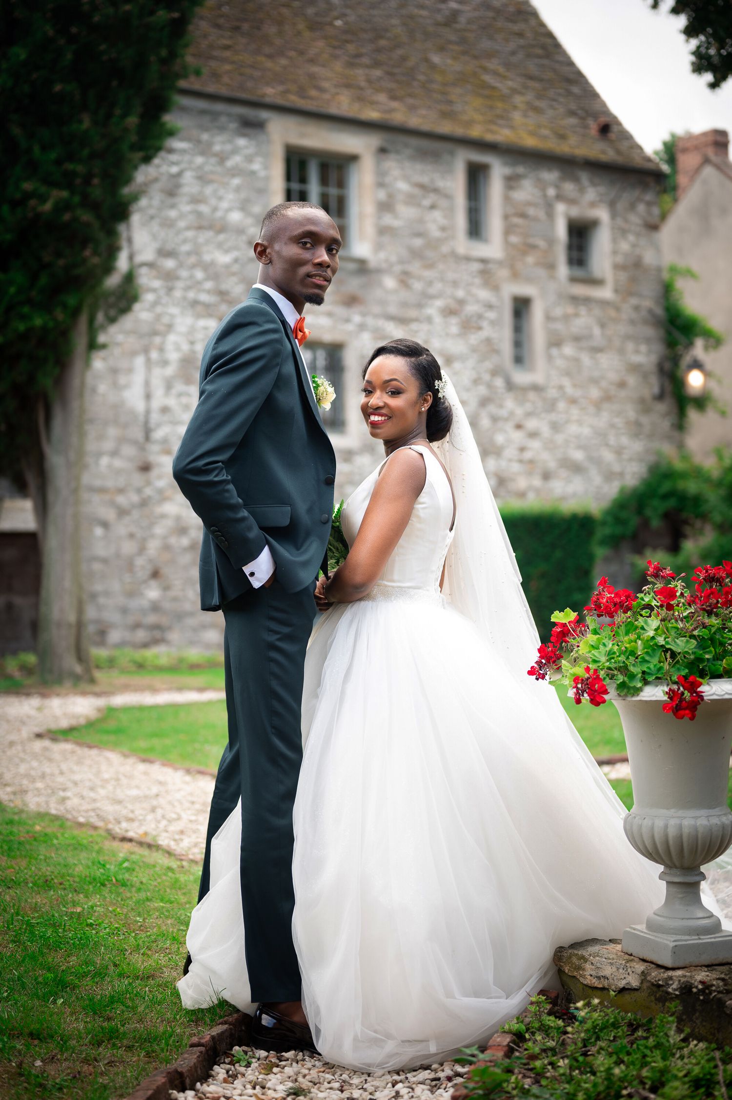 Bride and groom posing in front of a historic stone building with red flowers in the garden.