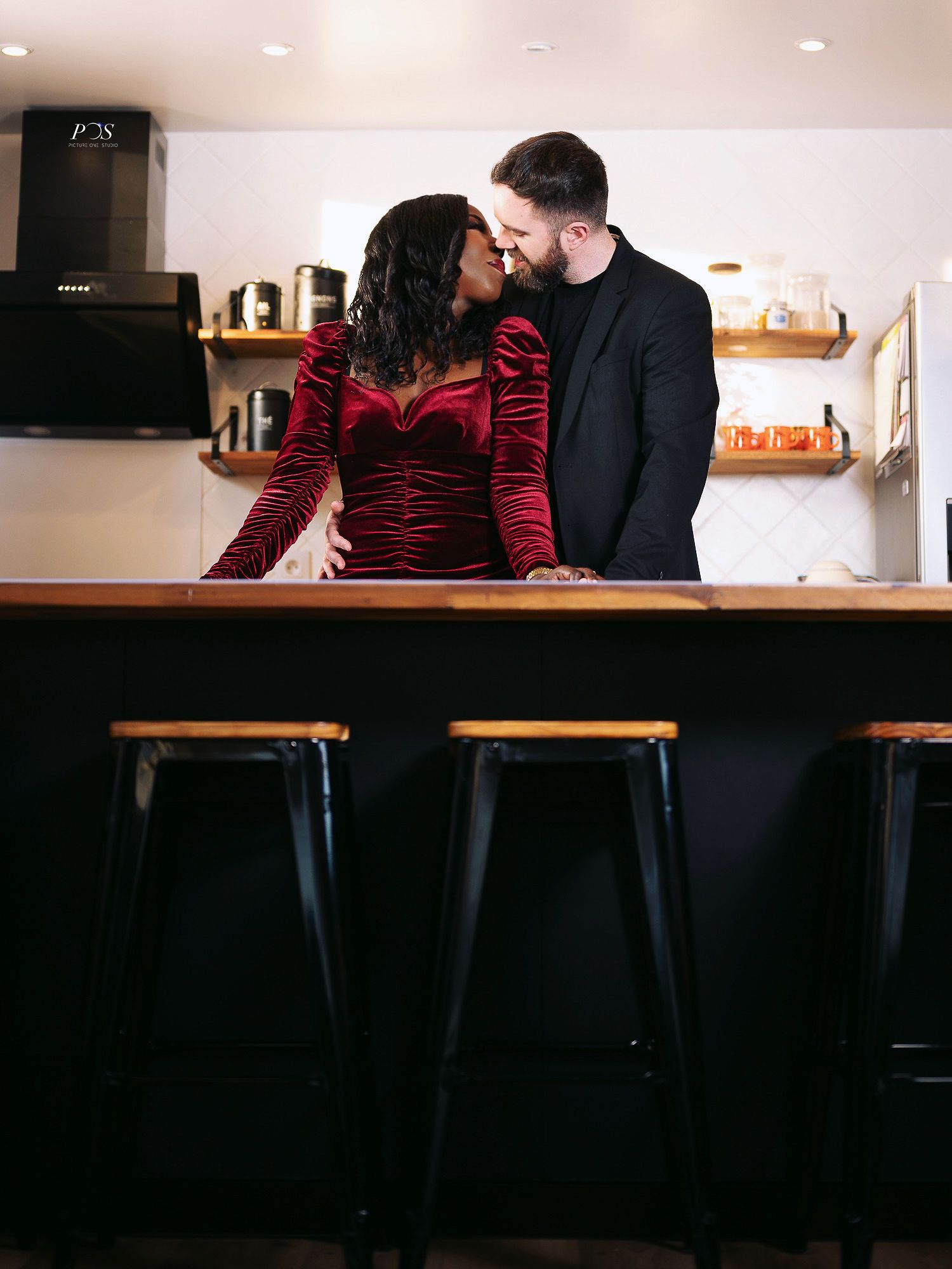 Couple sharing a romantic kiss over a kitchen bar counter with black stools in the foreground.