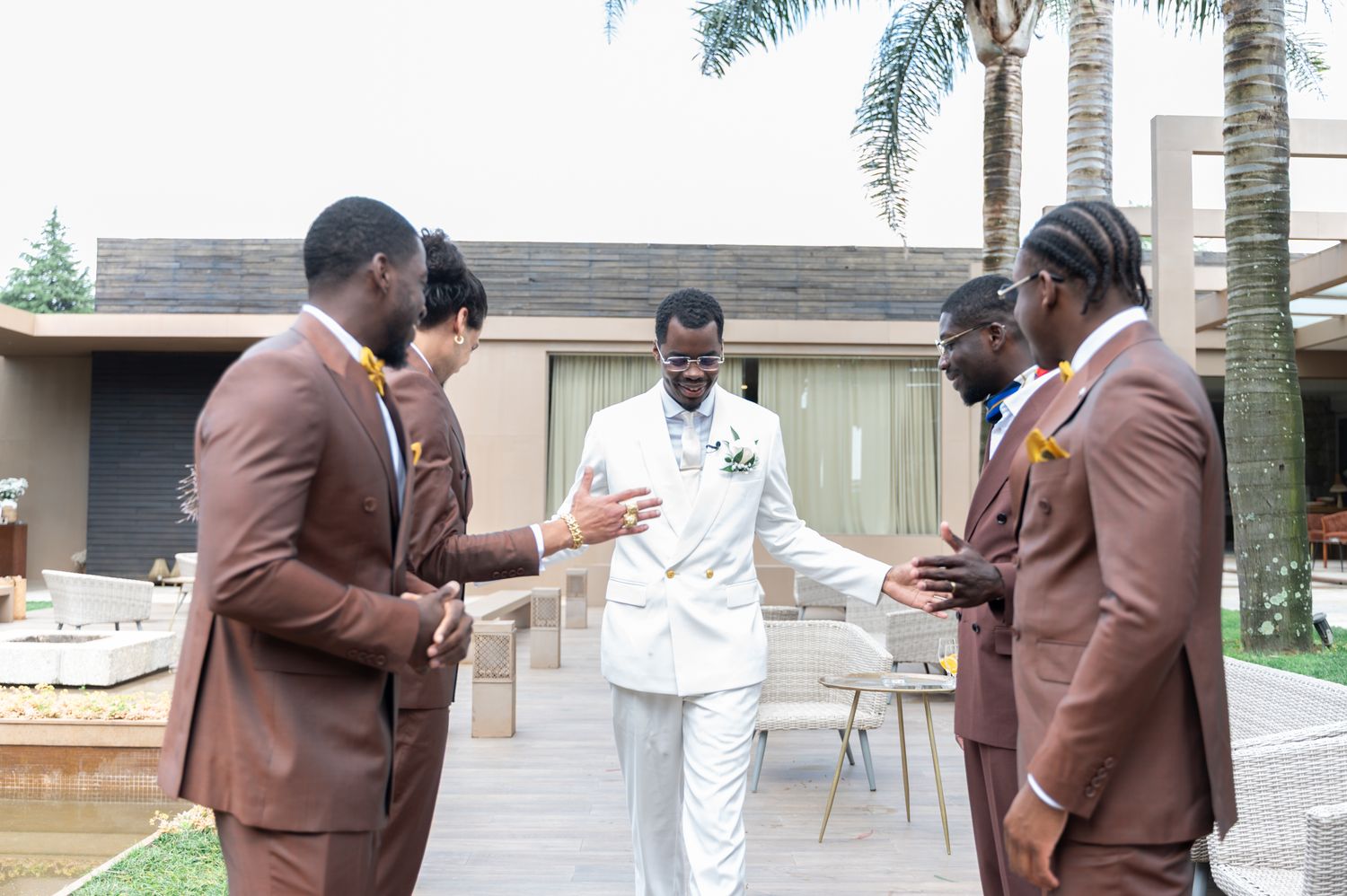 Groomsmen in brown suits surrounding groom in white suit at outdoor wedding venue.