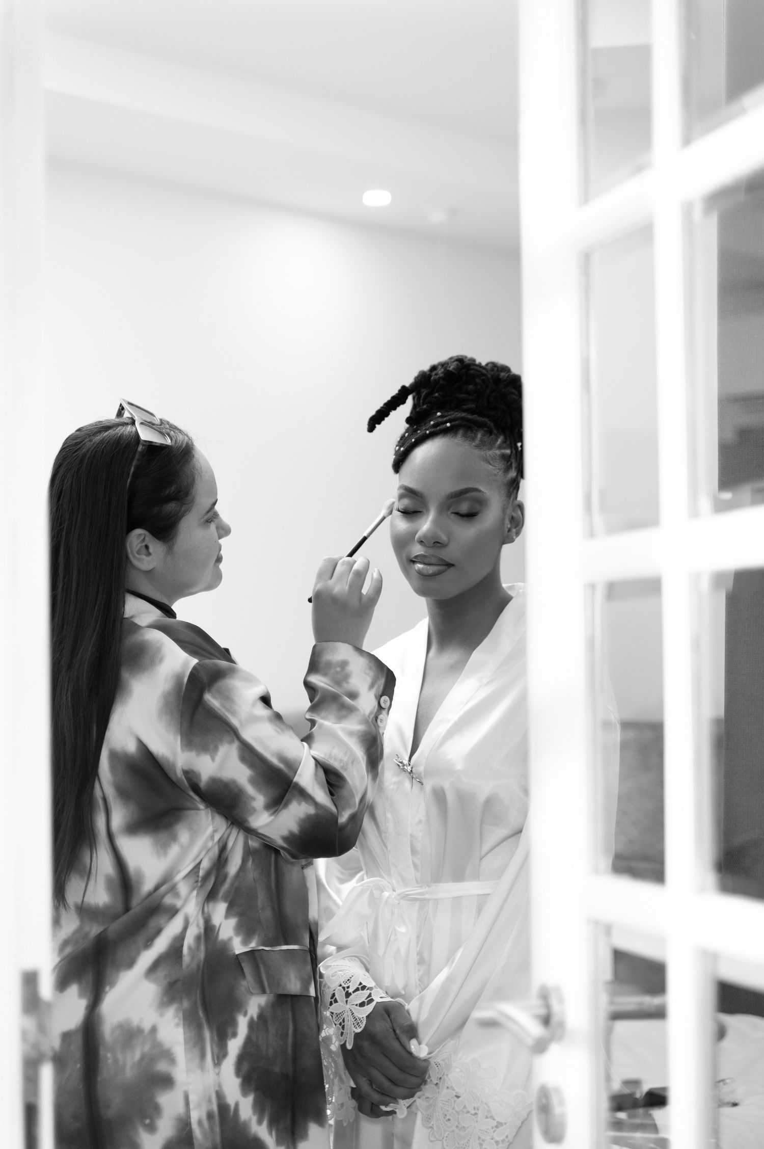 Black and white photo of makeup artist applying cosmetics to a bride in a white robe.