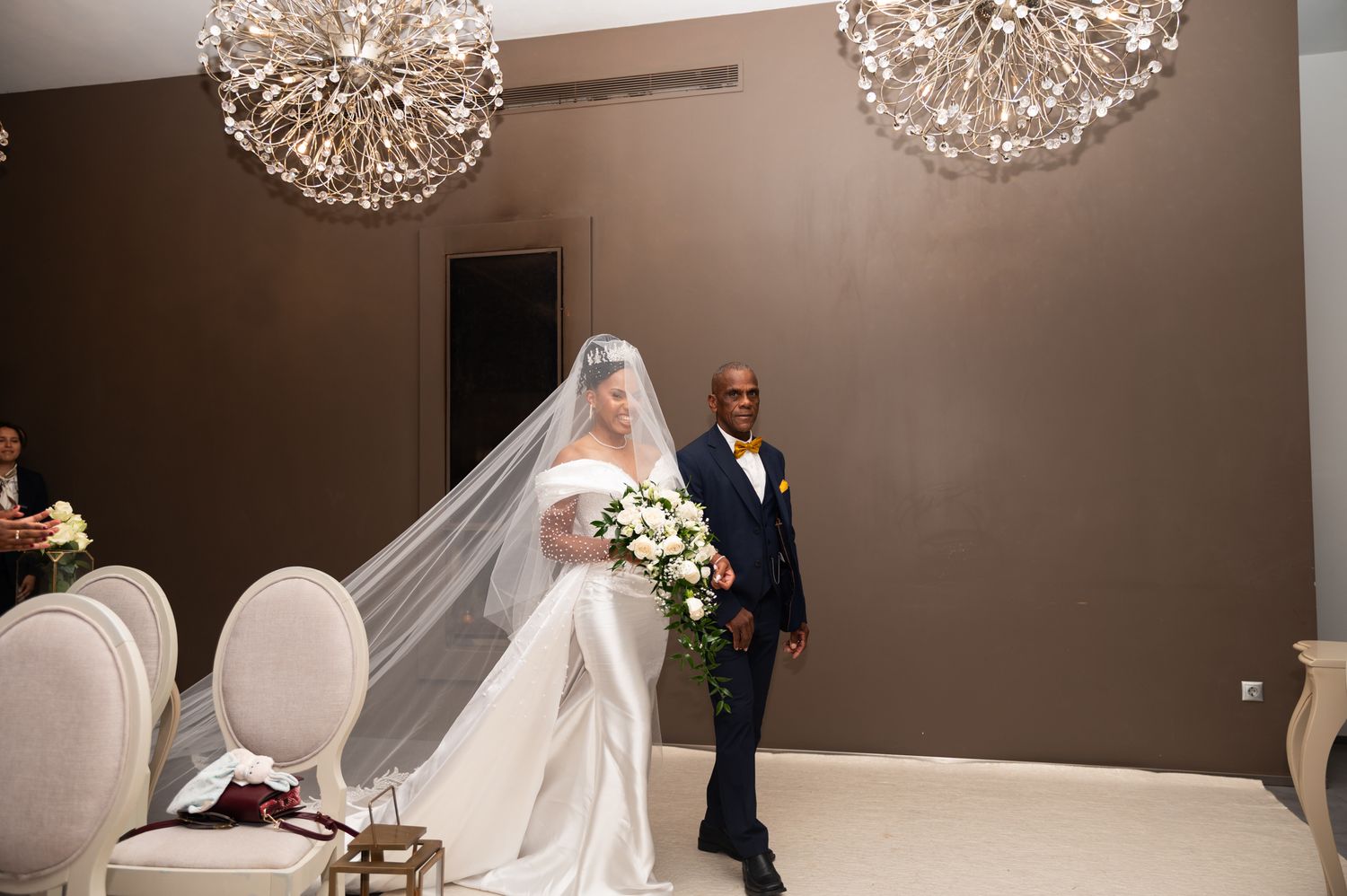 Bride in elegant white gown with long veil holds bouquet beside groom in navy suit indoors.