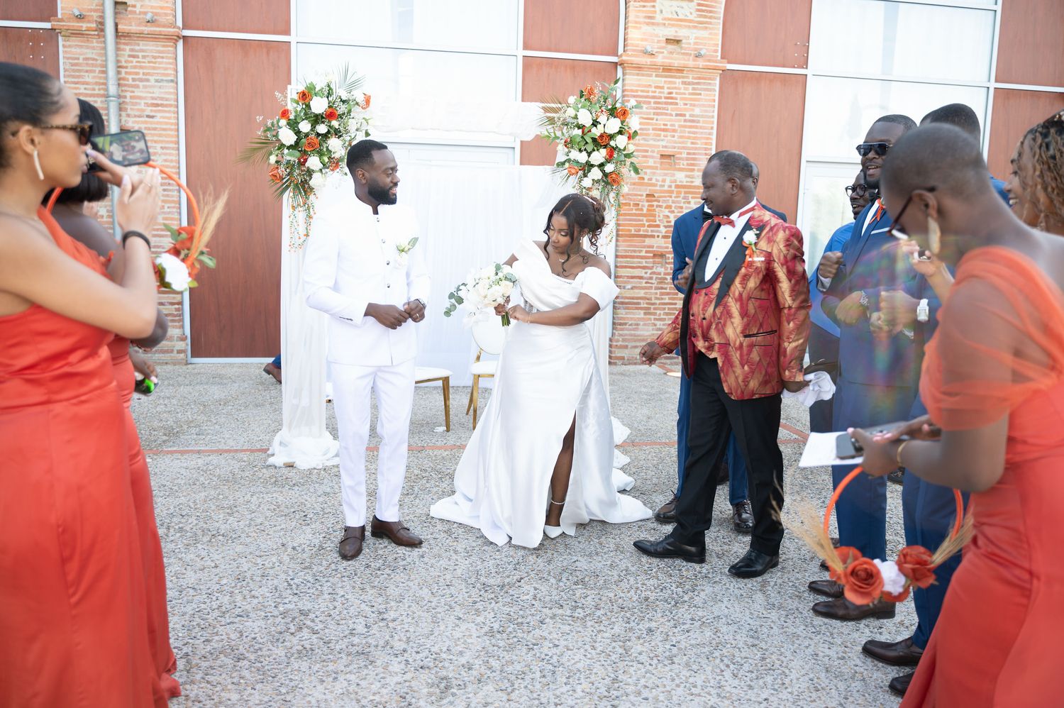 Wedding ceremony with bride and groom in white surrounded by guests in orange attire outdoors.