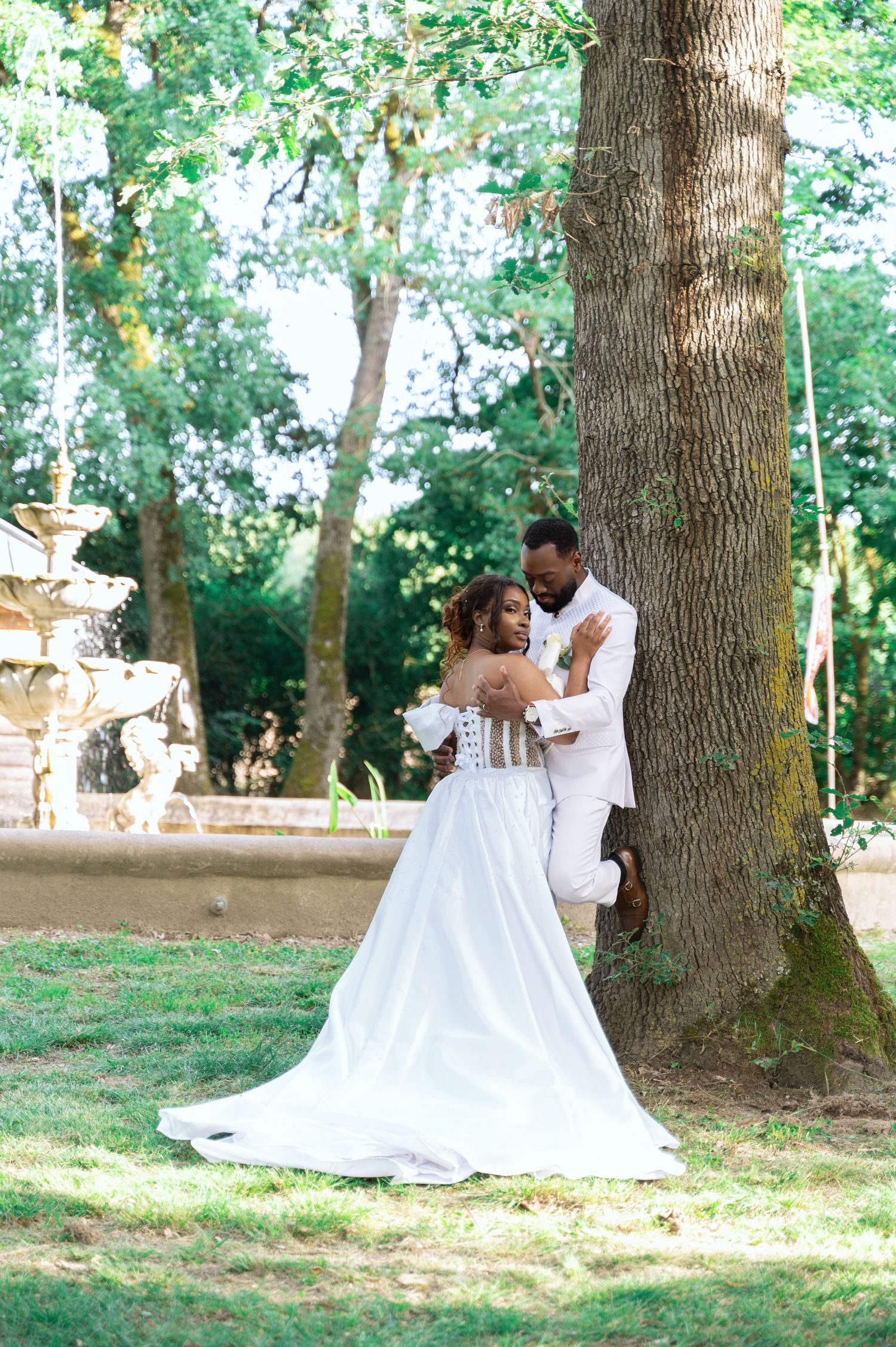 Groom in white suit embraces bride in strapless gown near large tree with fountain backdrop outdoors.