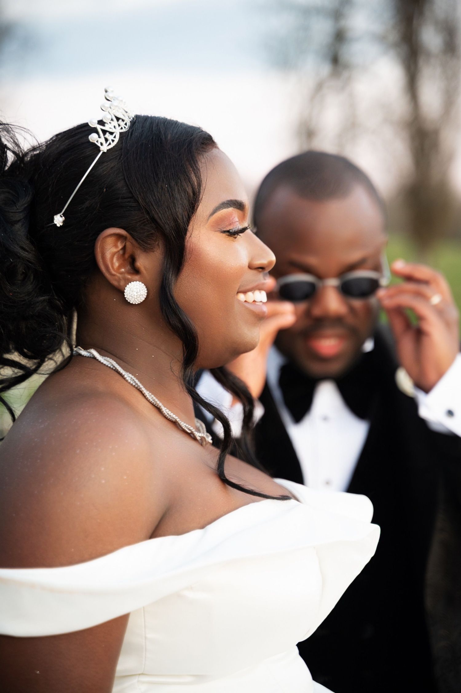 A glowing bride in an off-shoulder white gown smiles as her groom in a tuxedo playfully adjusts his sunglasses.