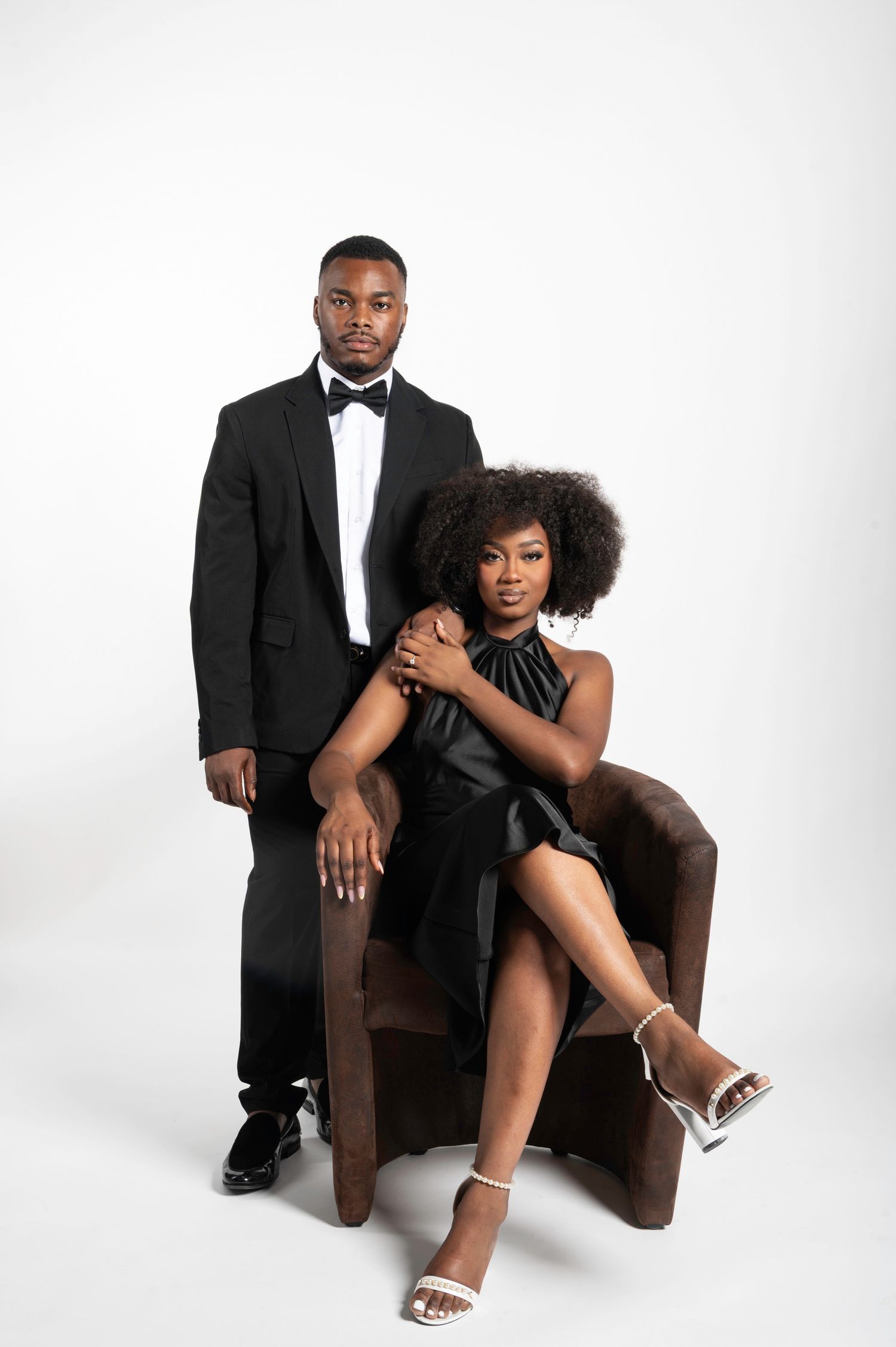 Elegant Black couple posing in formal attire, man in tuxedo standing behind woman in black dress seated in brown chair.