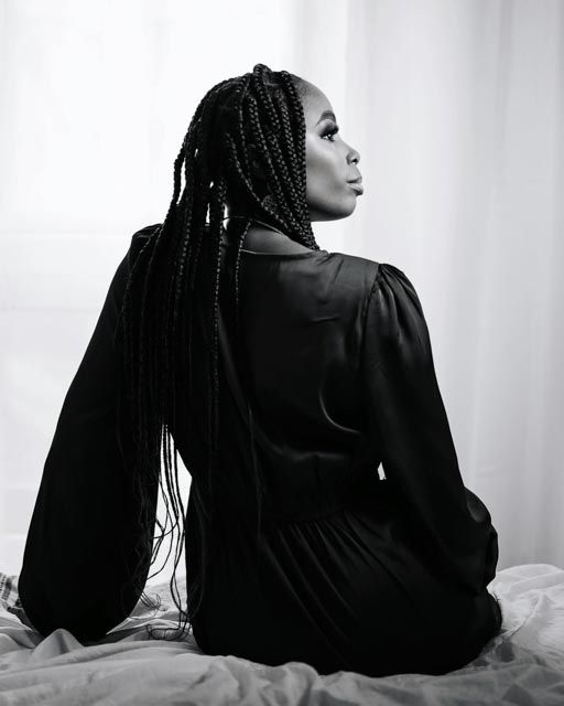 Black and white portrait of a woman with long box braids wearing a black satin dress, seated gracefully on a bed.