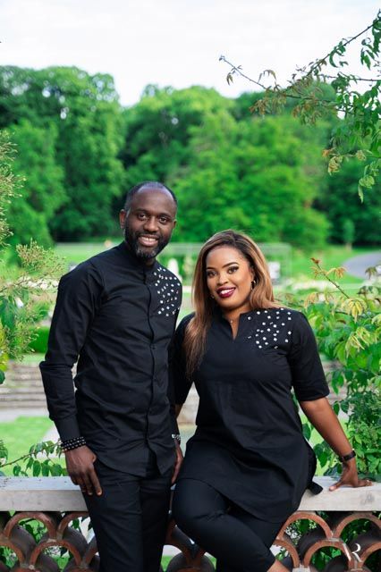 A couple dressed in matching black outfits with pearl accents pose together outdoors in a lush green garden setting.