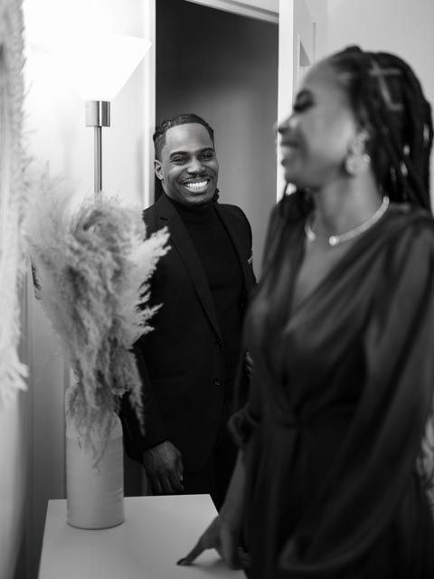 Black and white photo of a smiling man in a black suit laughing with a woman wearing braids in an elegant interior.