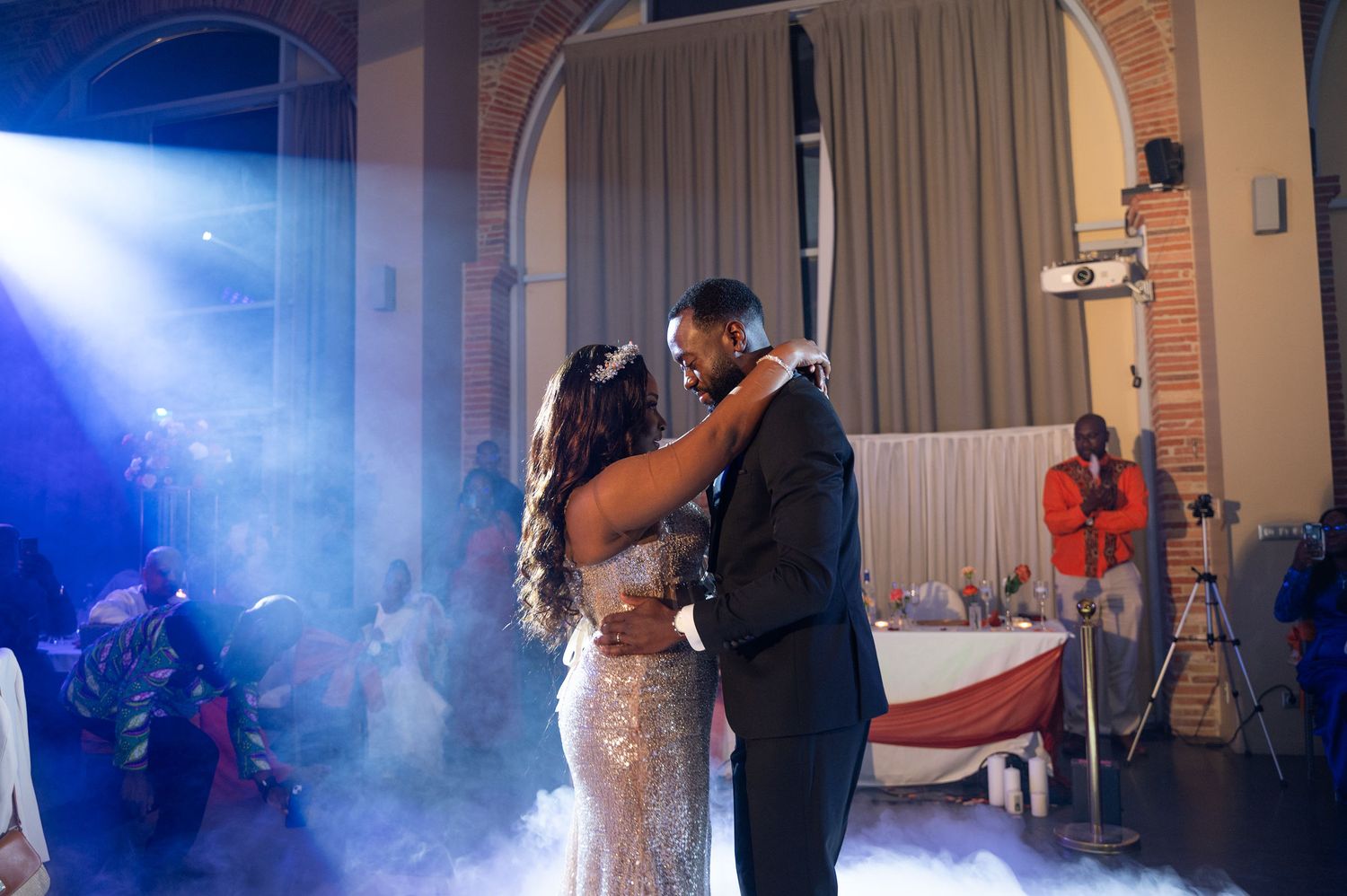 Couple shares romantic first dance on a fog-covered monogrammed dance floor with blue spotlights and wedding guests.
