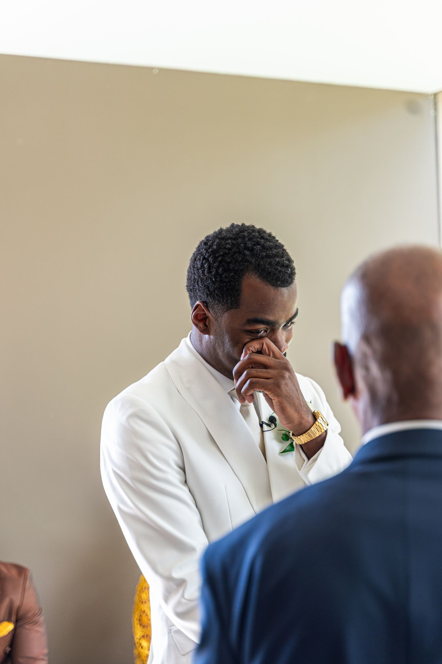 Emotional groom in white suit wipes tears during his wedding ceremony while standing at the altar.