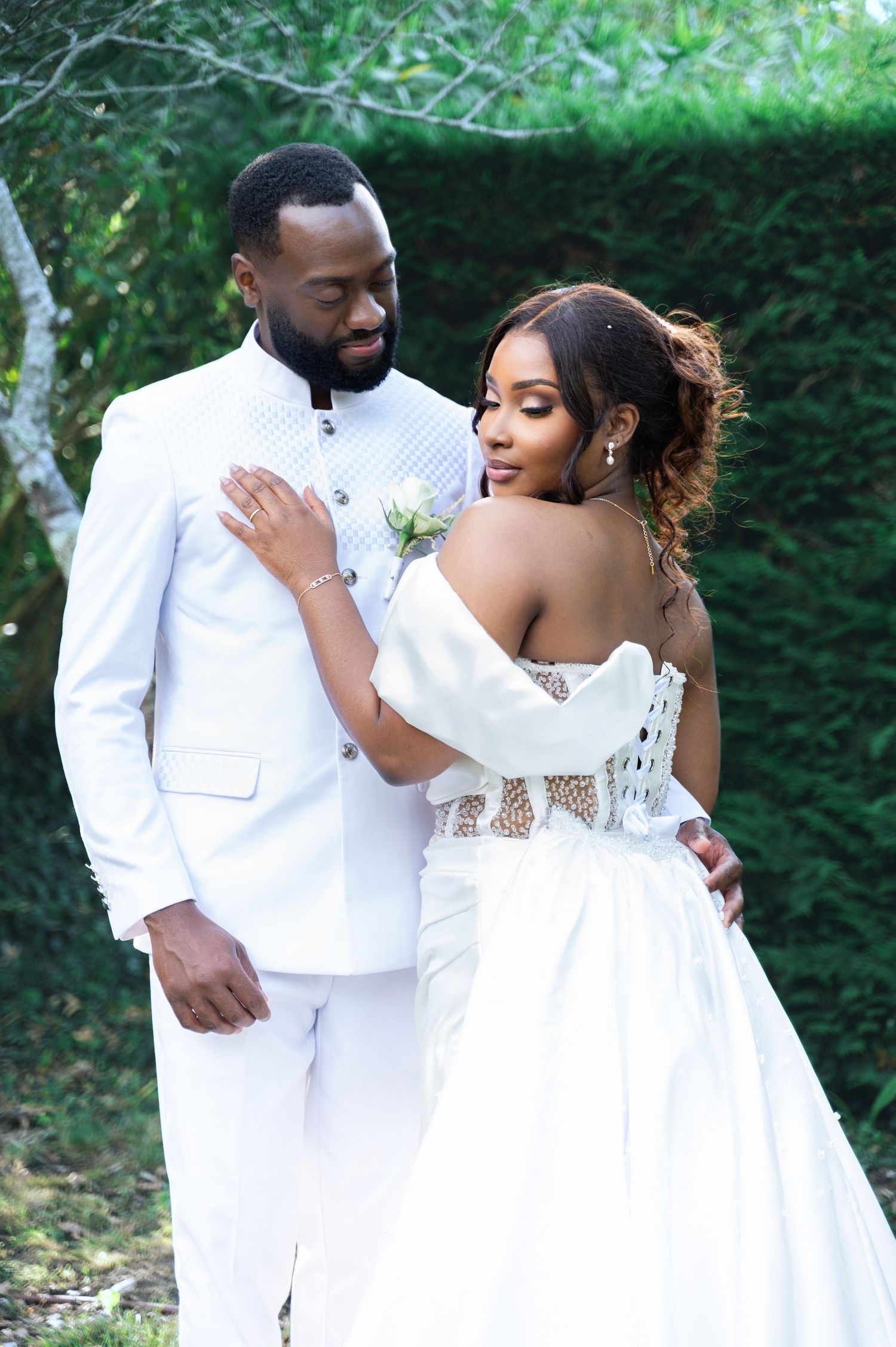 A Black bride and groom in white wedding attire share a tender embrace surrounded by lush greenery.