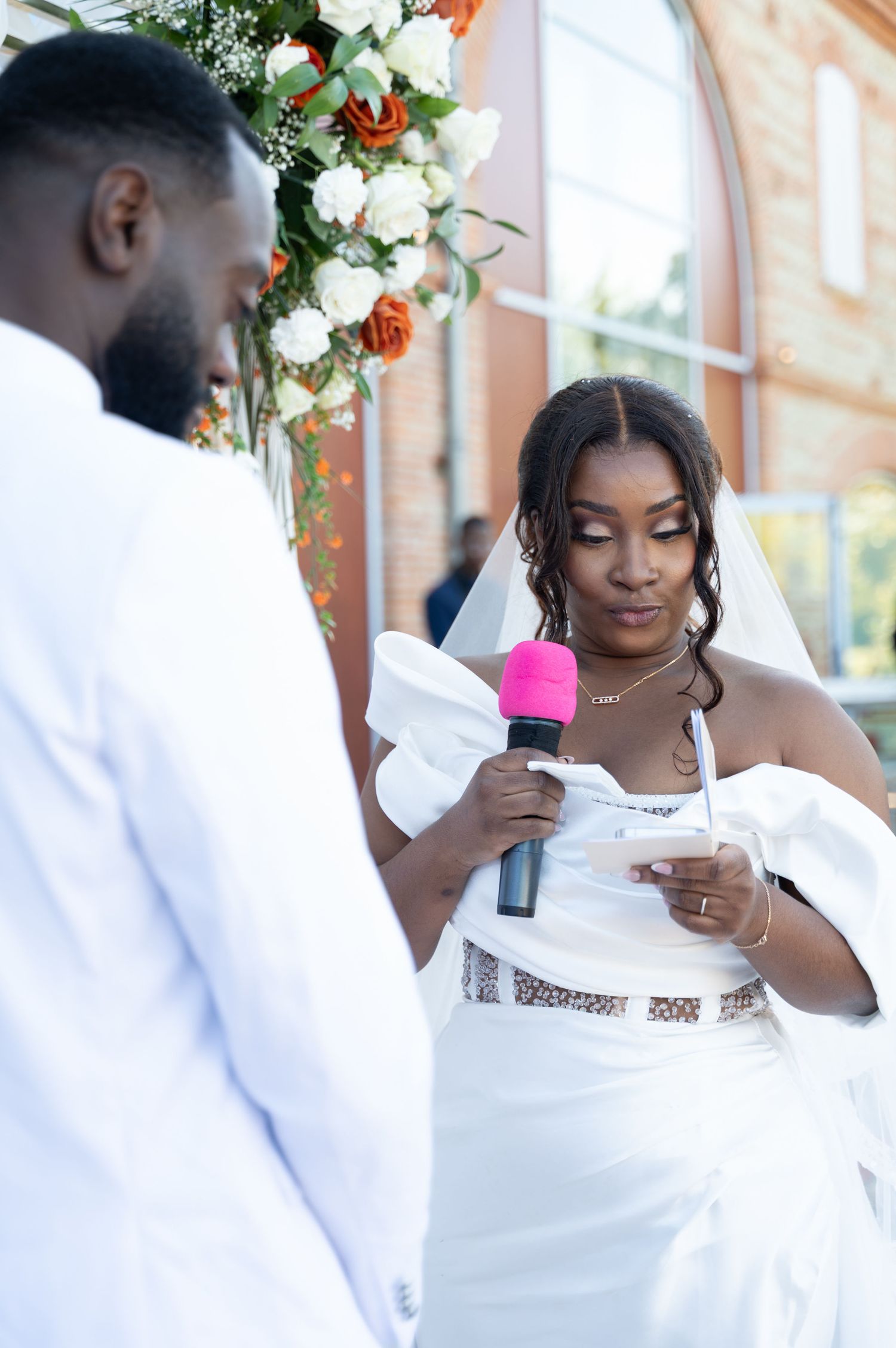 Bride in white gown reads vows into pink microphone at outdoor wedding ceremony.