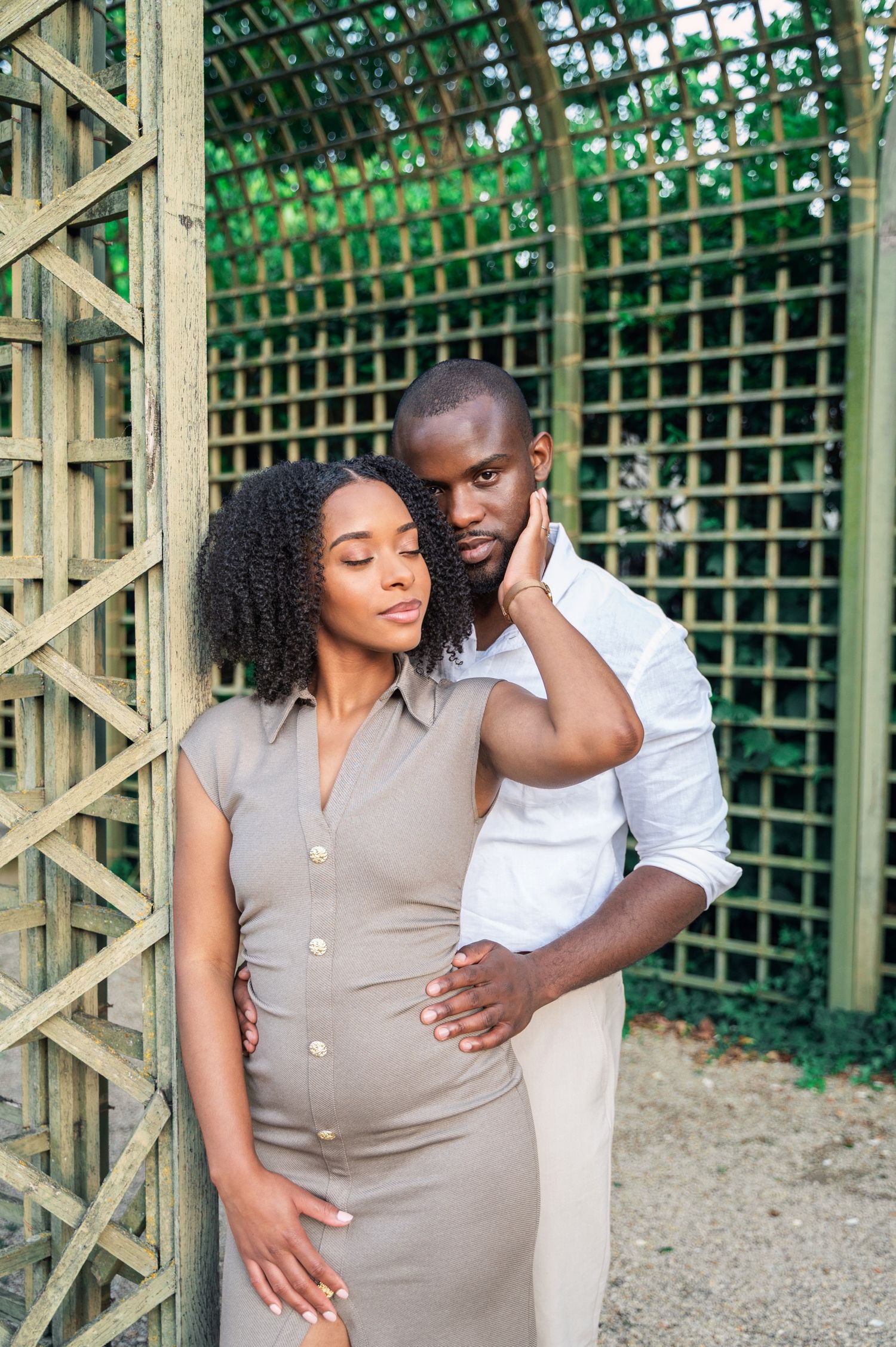 A Black couple shares a tender embrace leaning against a wooden lattice wall in a lush garden setting.