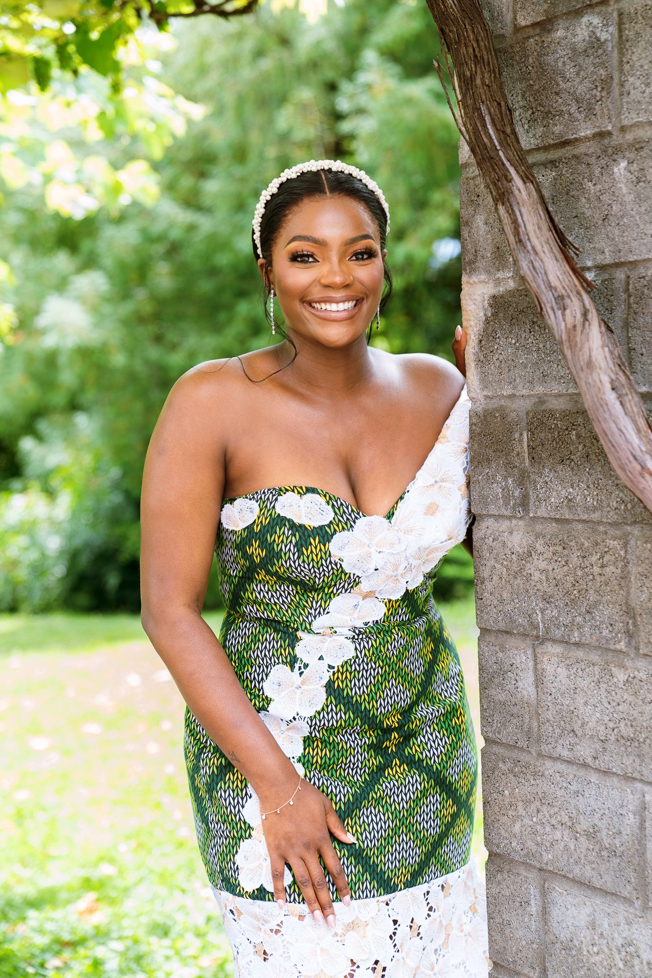 Smiling bride in strapless green African print dress with white floral detail posing by stone wall outdoors.