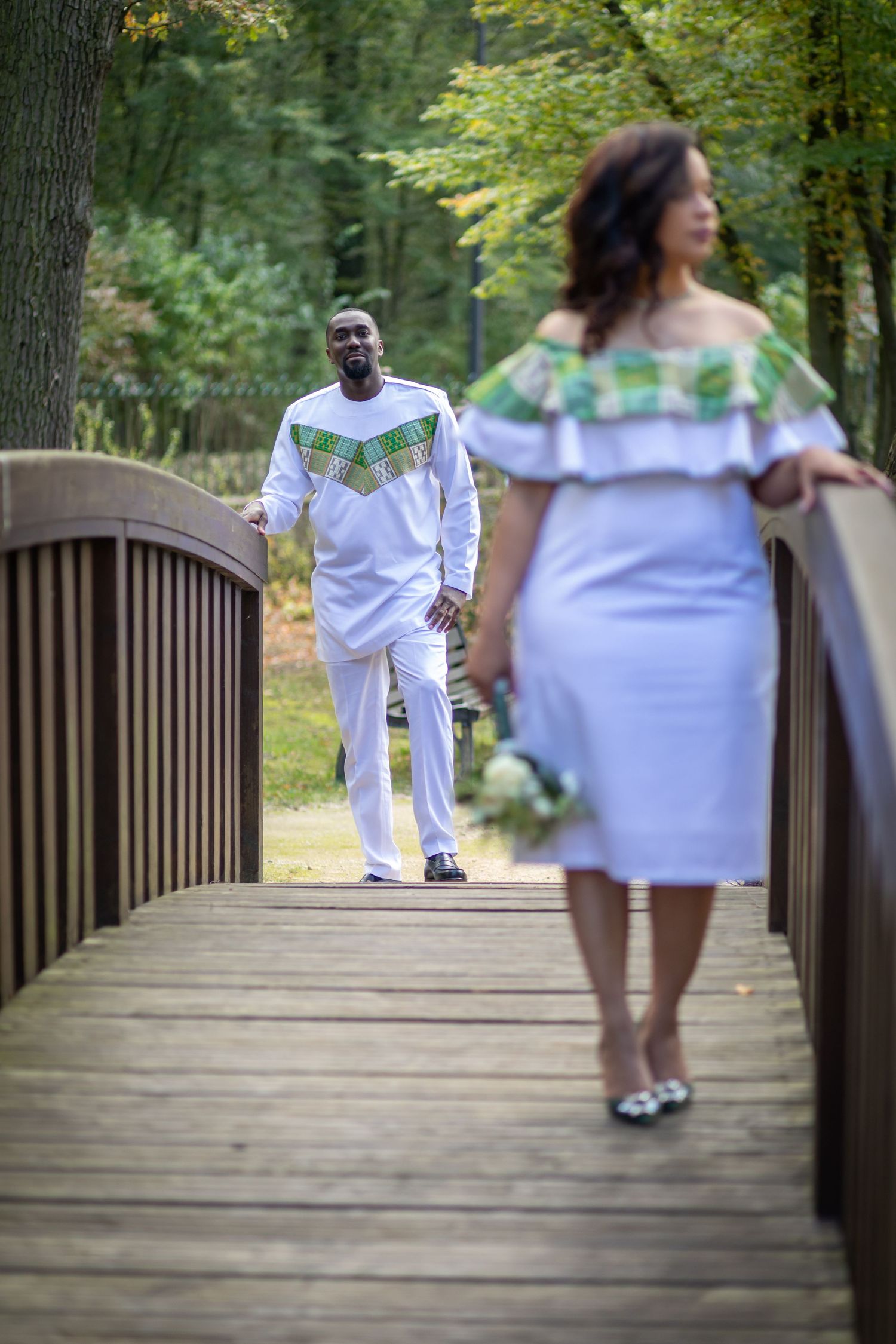 Couple in African print white outfits standing on wooden bridge in lush green park setting.