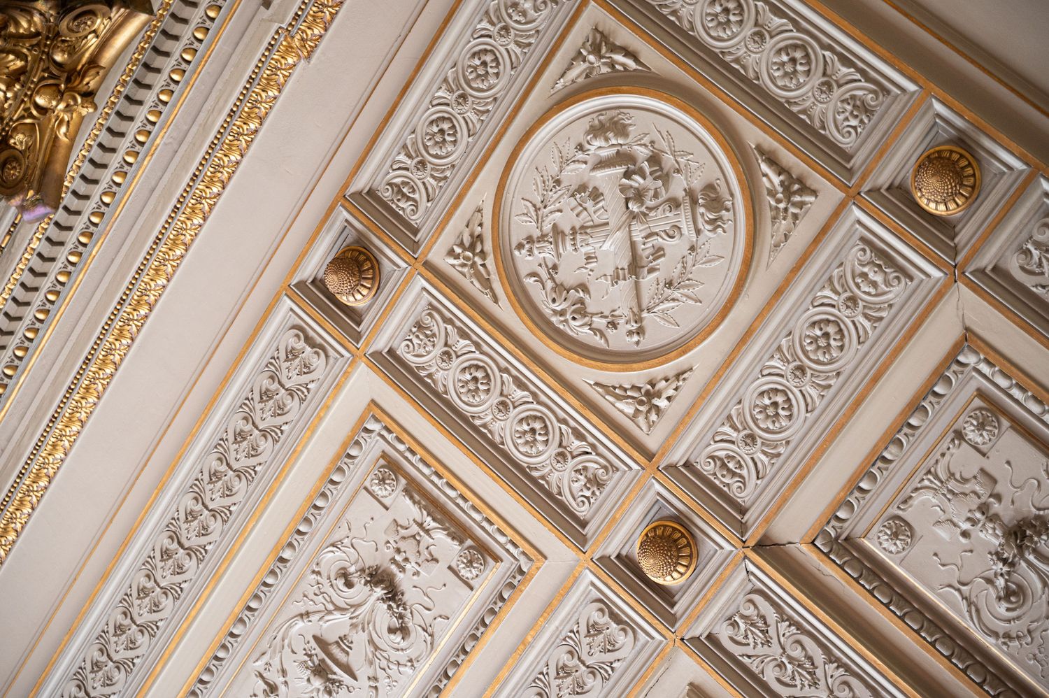 Ornate gilded ceiling with intricate white plasterwork reliefs and decorative moldings in a grand venue.