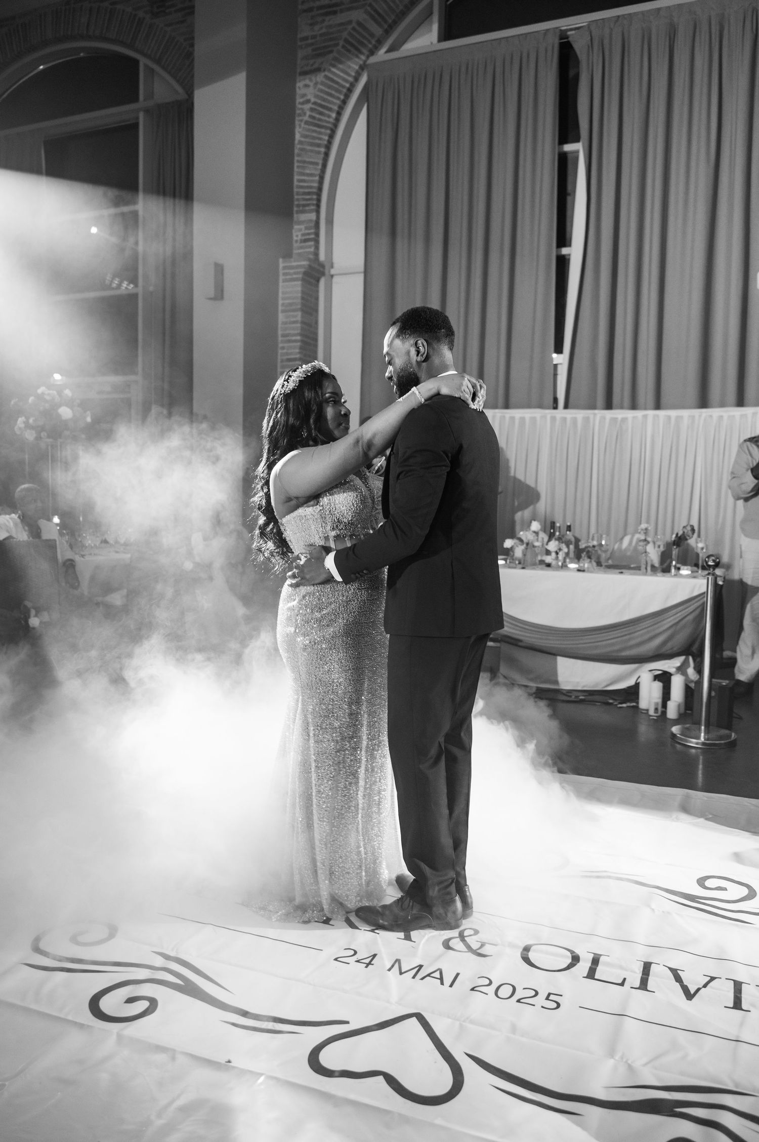 Black and white photo of bride and groom dancing on monogrammed floor reading 24 Mai 2025 with dreamy fog effects.