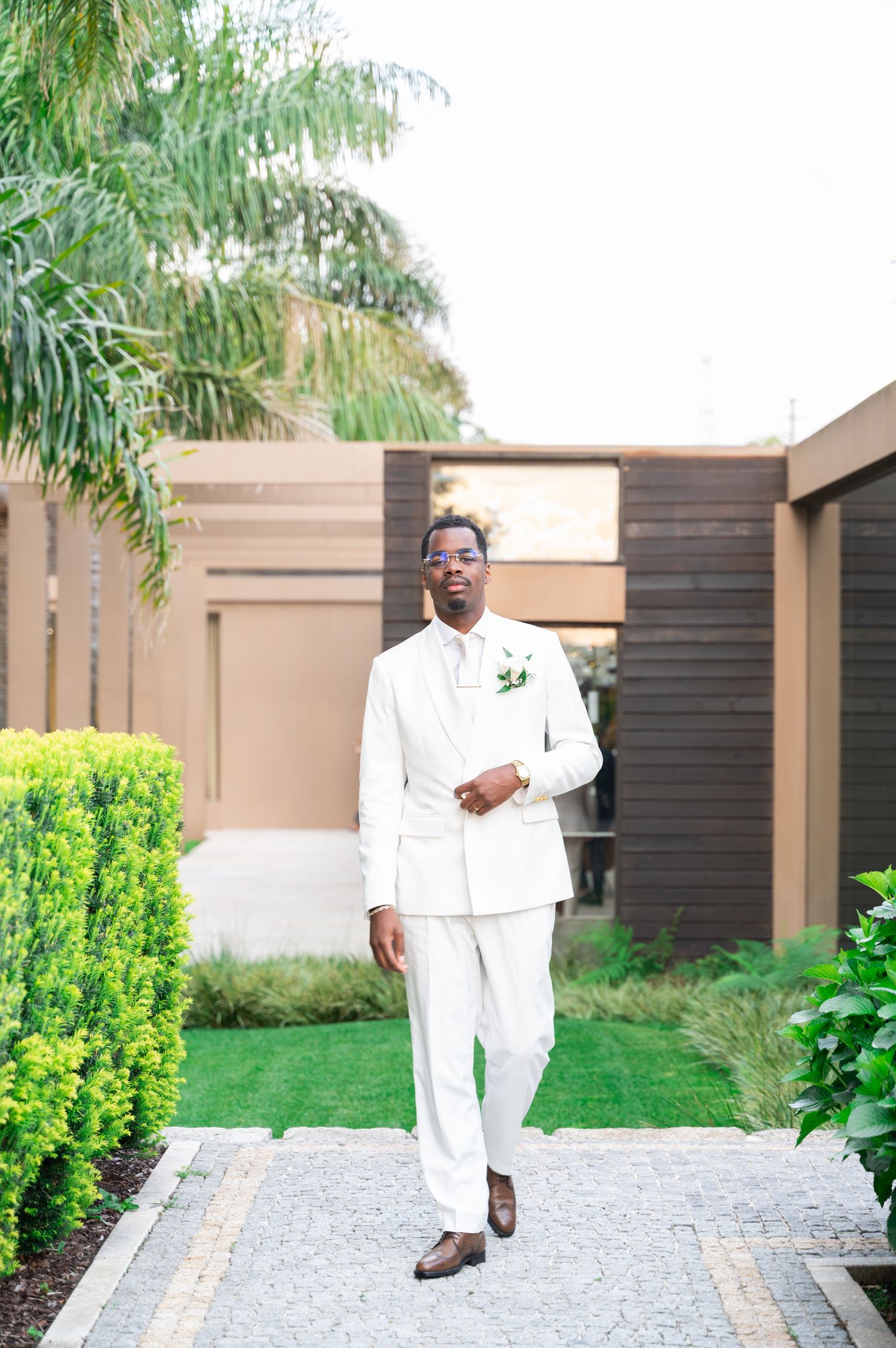 Dapper groom in white suit with boutonniere walks confidently through lush garden pathway.
