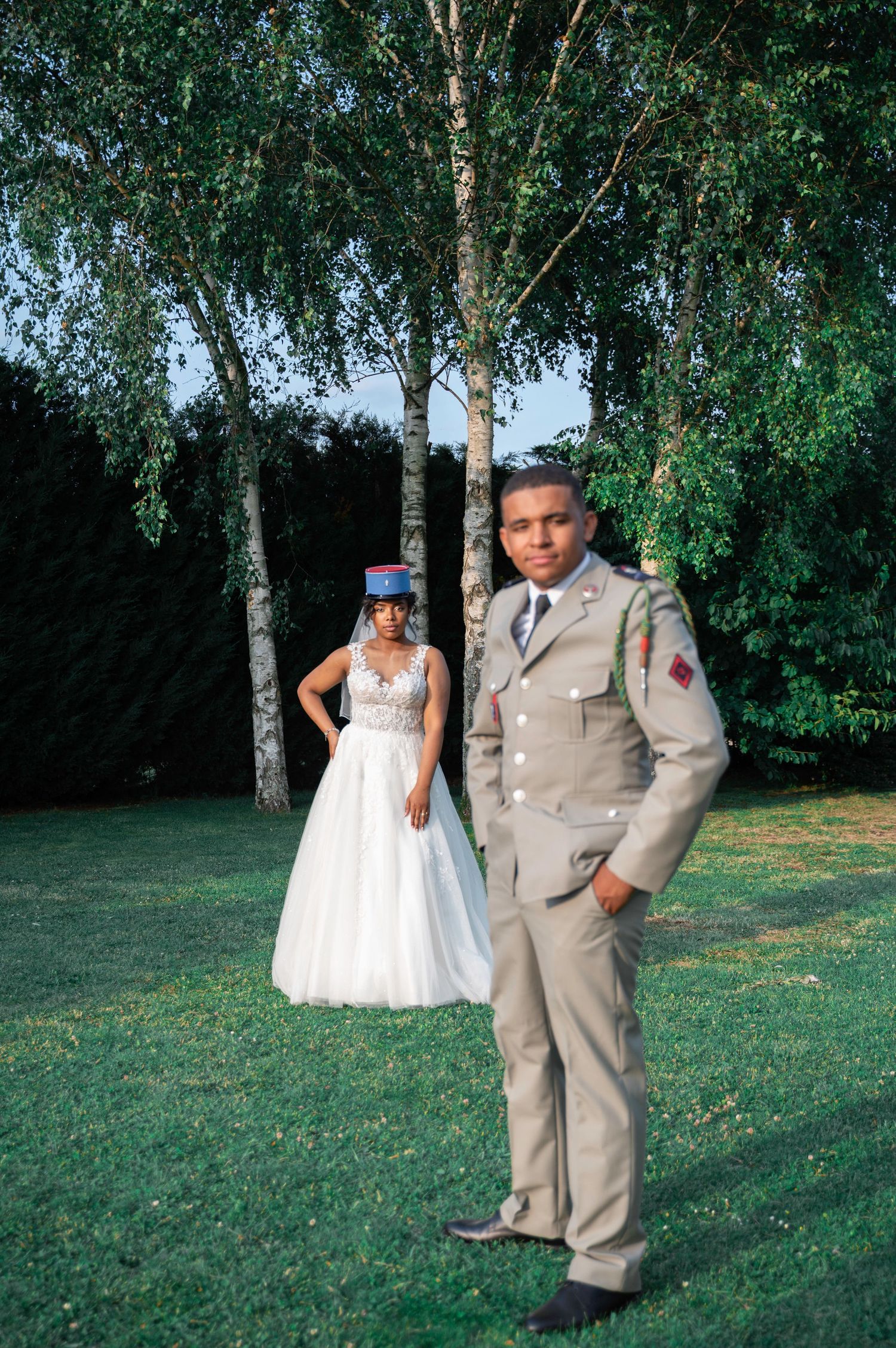 Military groom in tan uniform stands before bride in white gown among birch trees.