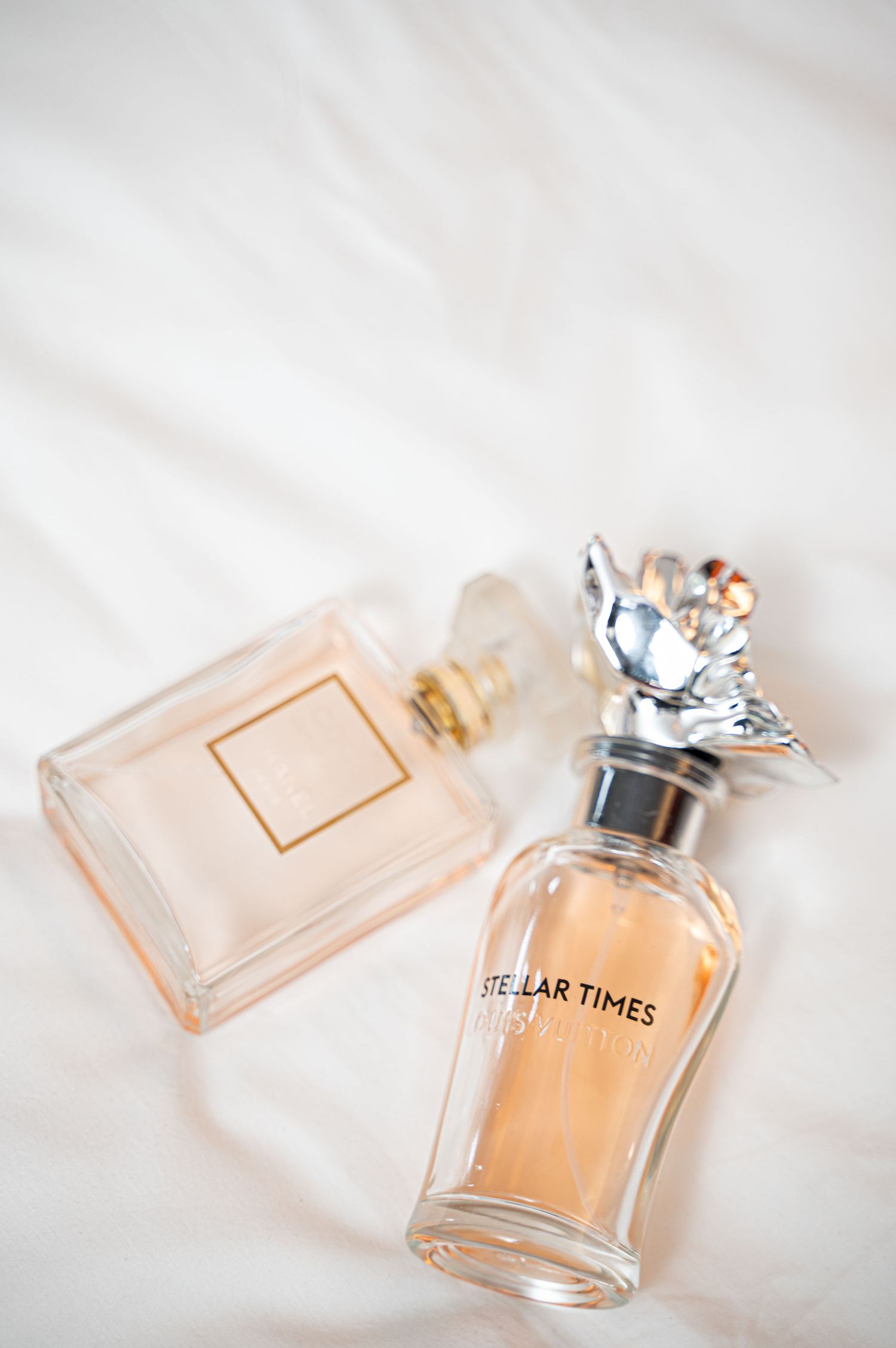 Two elegant perfume bottles, including Stellar Times fragrance, arranged on soft white fabric background.