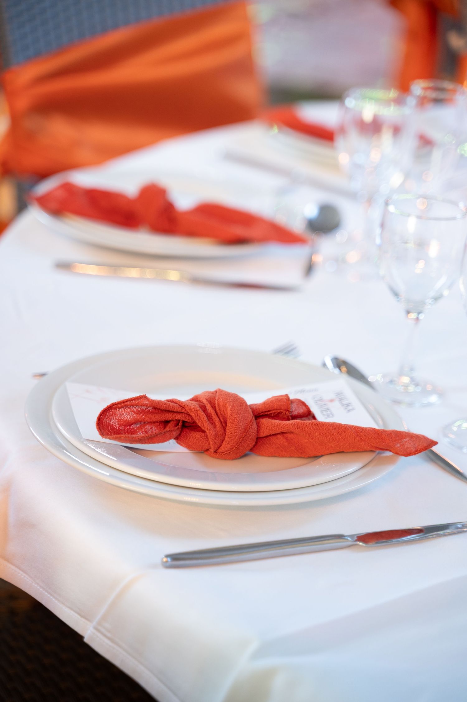 Elegant wedding table setting with red folded napkin on white plates, silverware, and crystal glasses.