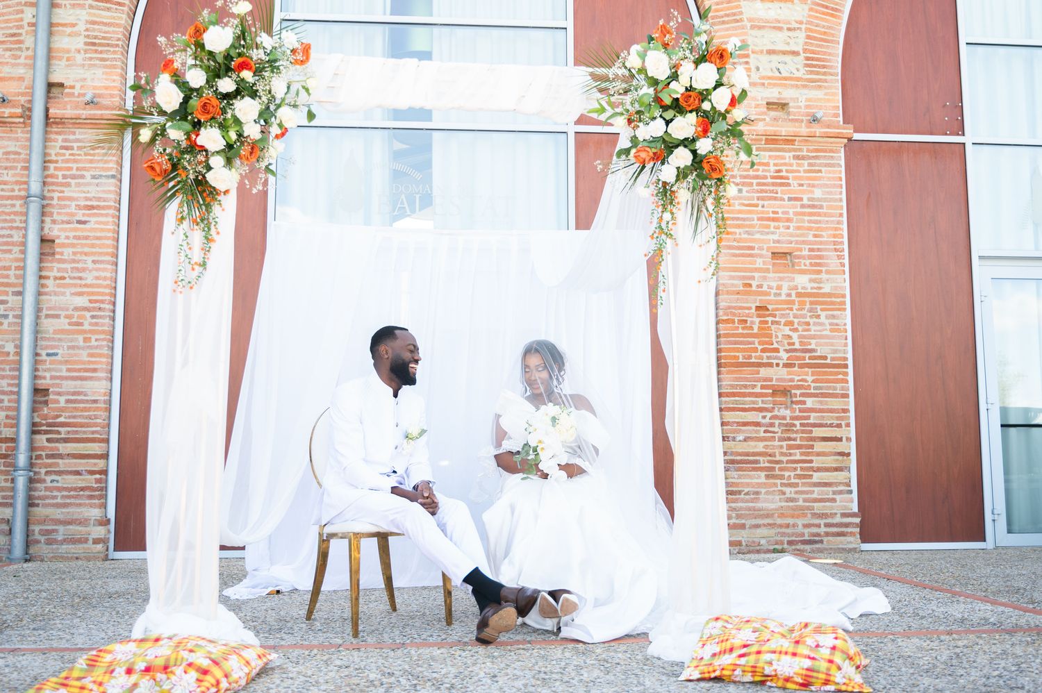 Bride and groom in white attire sit under floral arch with colorful African cushions.