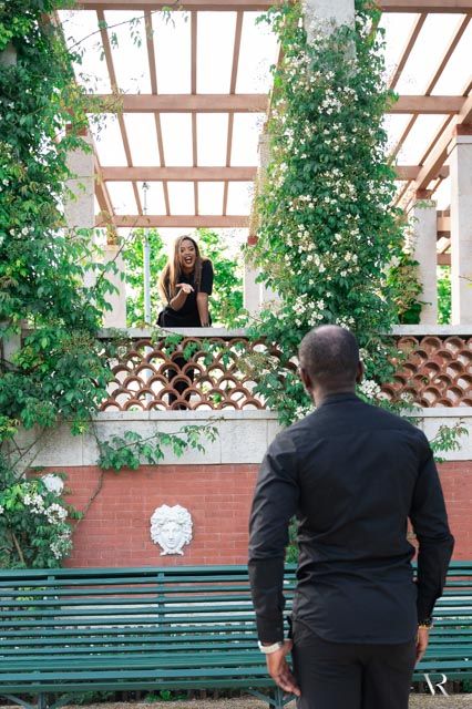 A man in black stands before a garden pergola where a woman smiles down at him amid lush greenery.