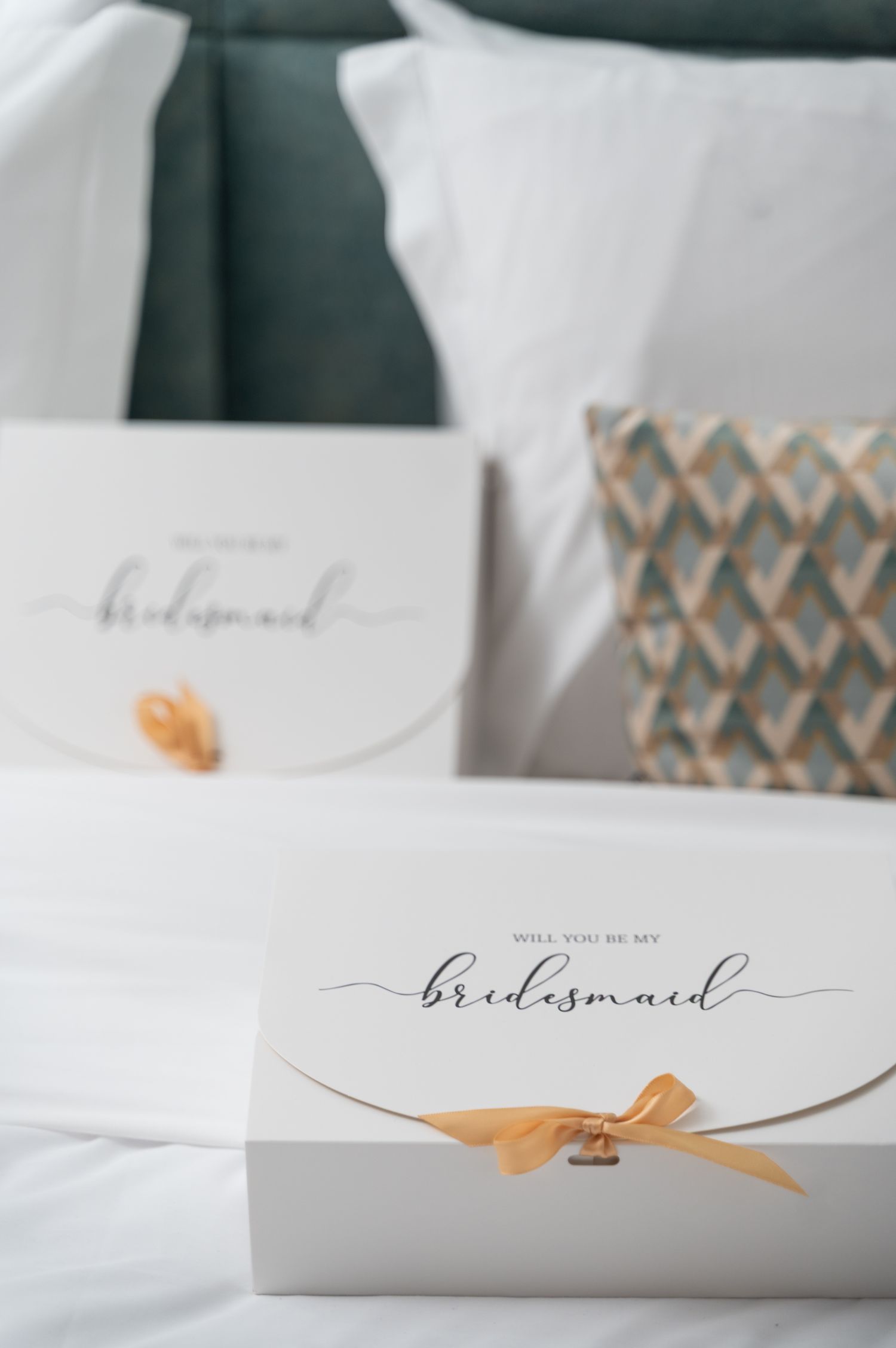 Two white bridesmaid proposal boxes with gold ribbon bows on white bedding.