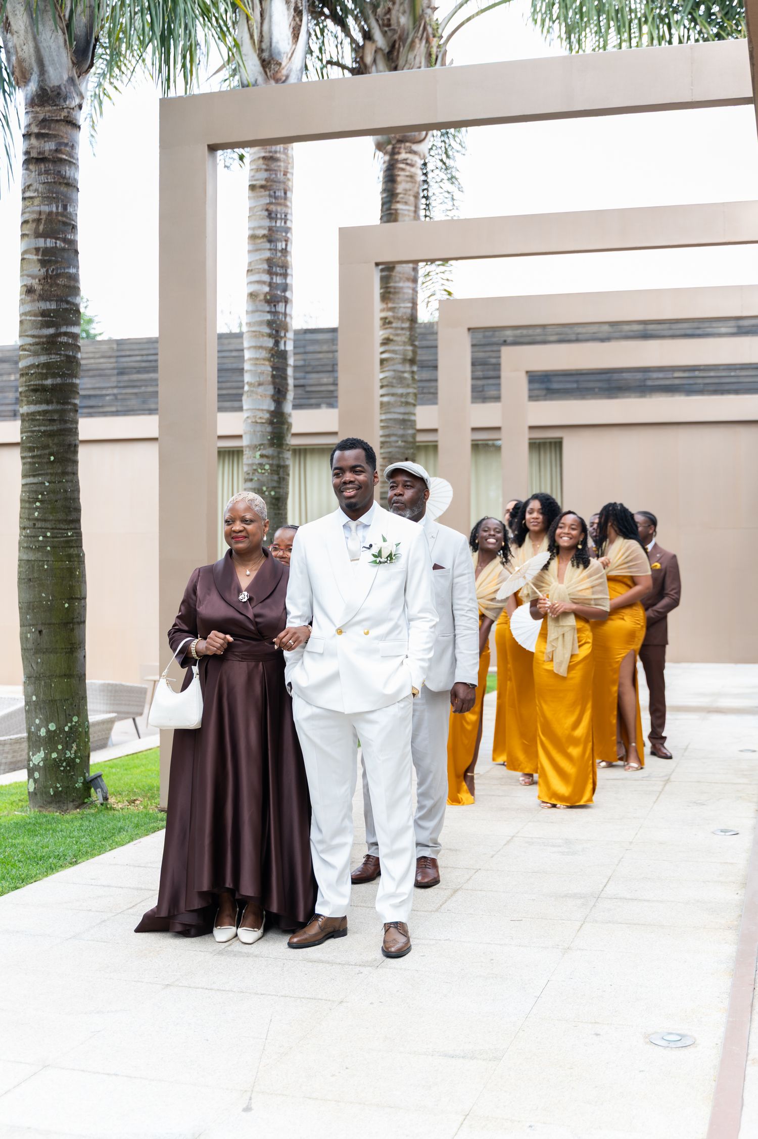 Groom in white suit escorts a woman in brown dress down an outdoor aisle with wedding party behind.