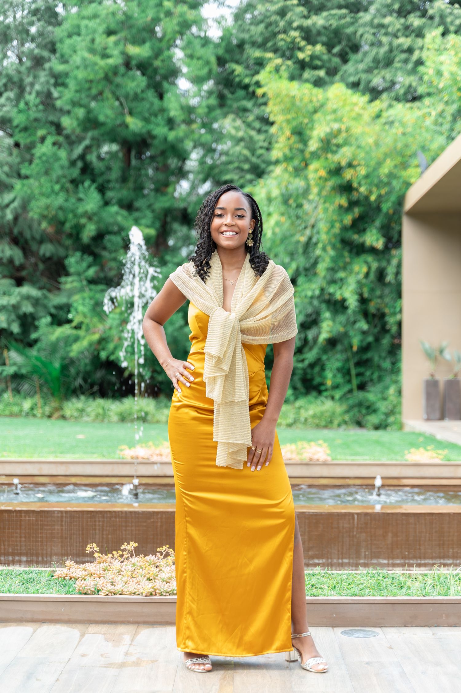 Smiling woman in mustard yellow gown with gold shawl poses confidently by lush garden fountain.