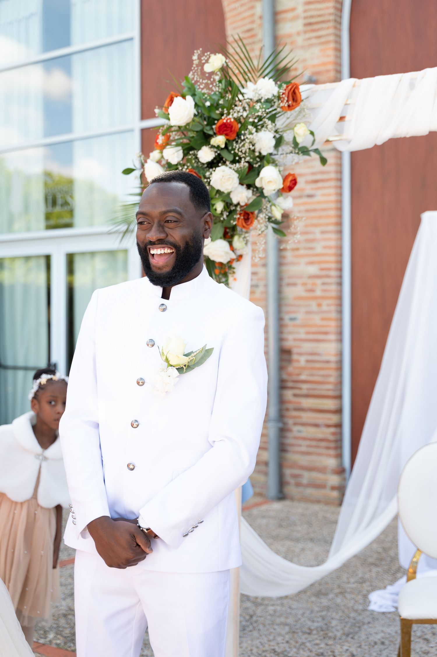 Smiling groom in white suit stands beneath floral arch at outdoor wedding ceremony.